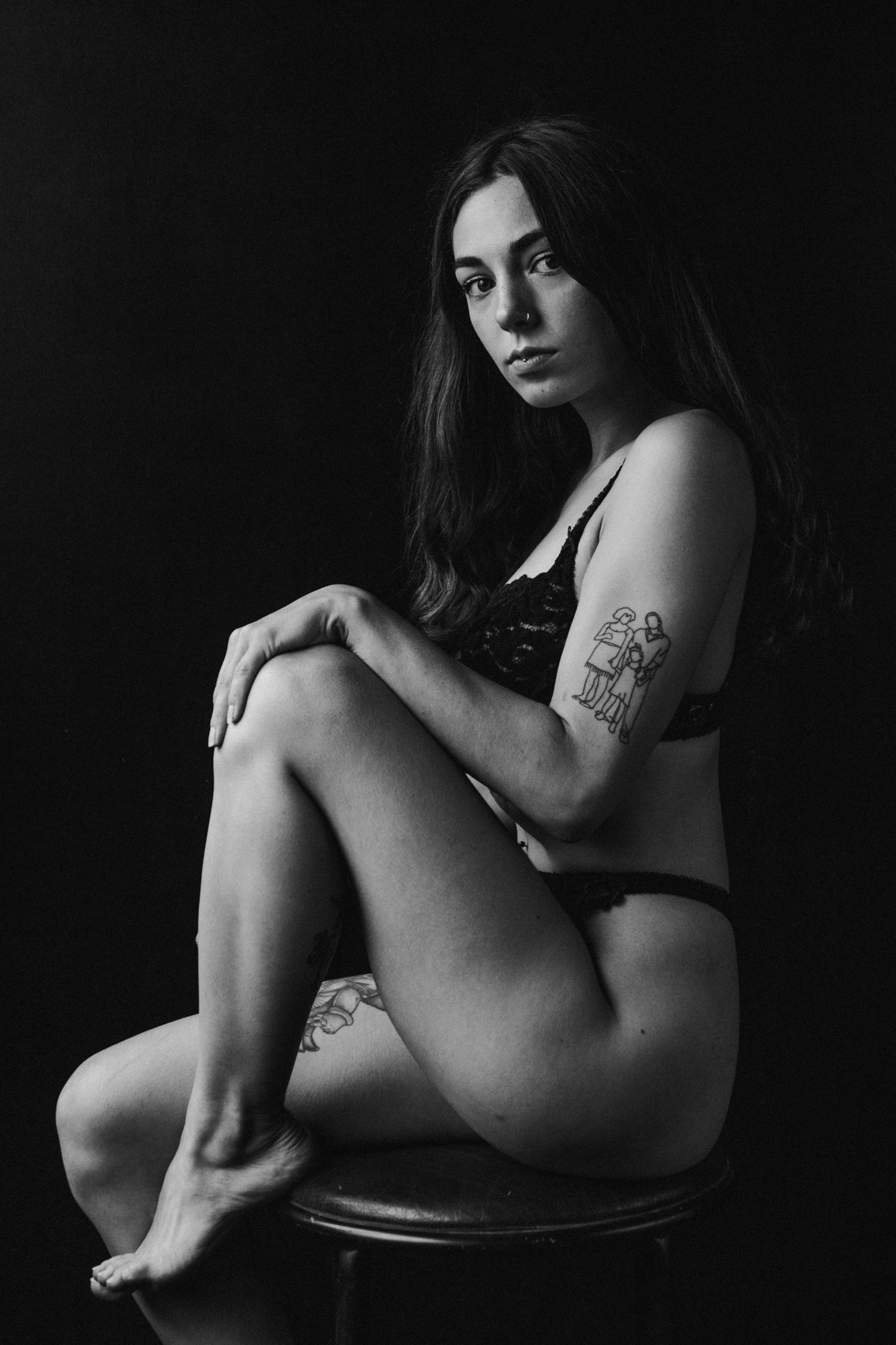 Black and white portrait of tattooed brunette woman in lingerie seated on stool.