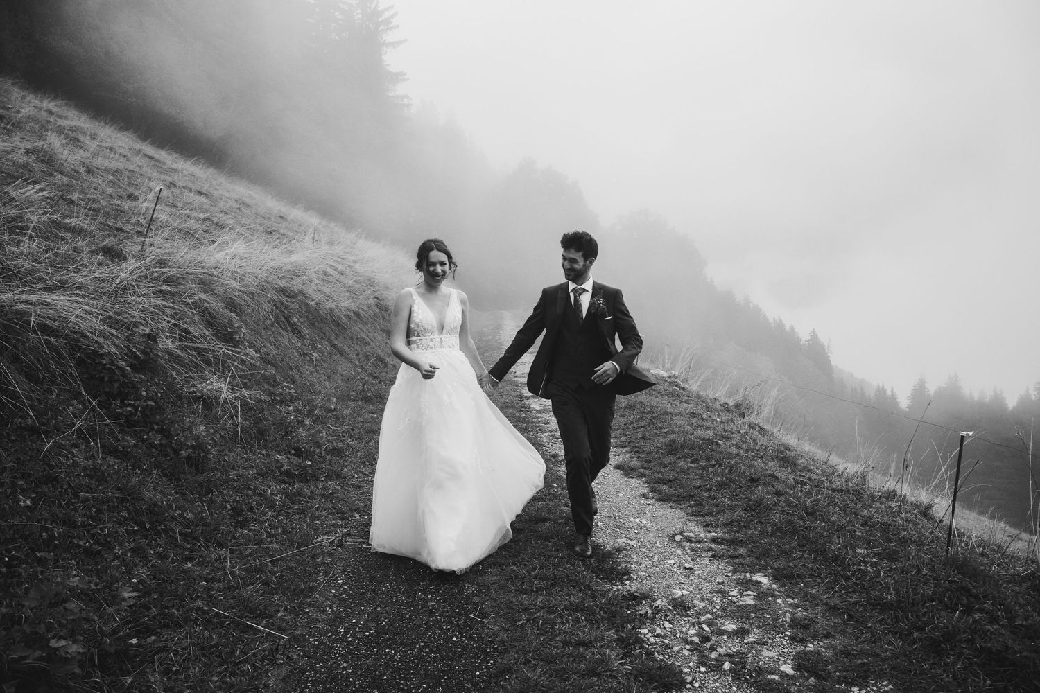 Black and white photo of newlyweds walking hand in hand along a misty mountain path in wedding attire.