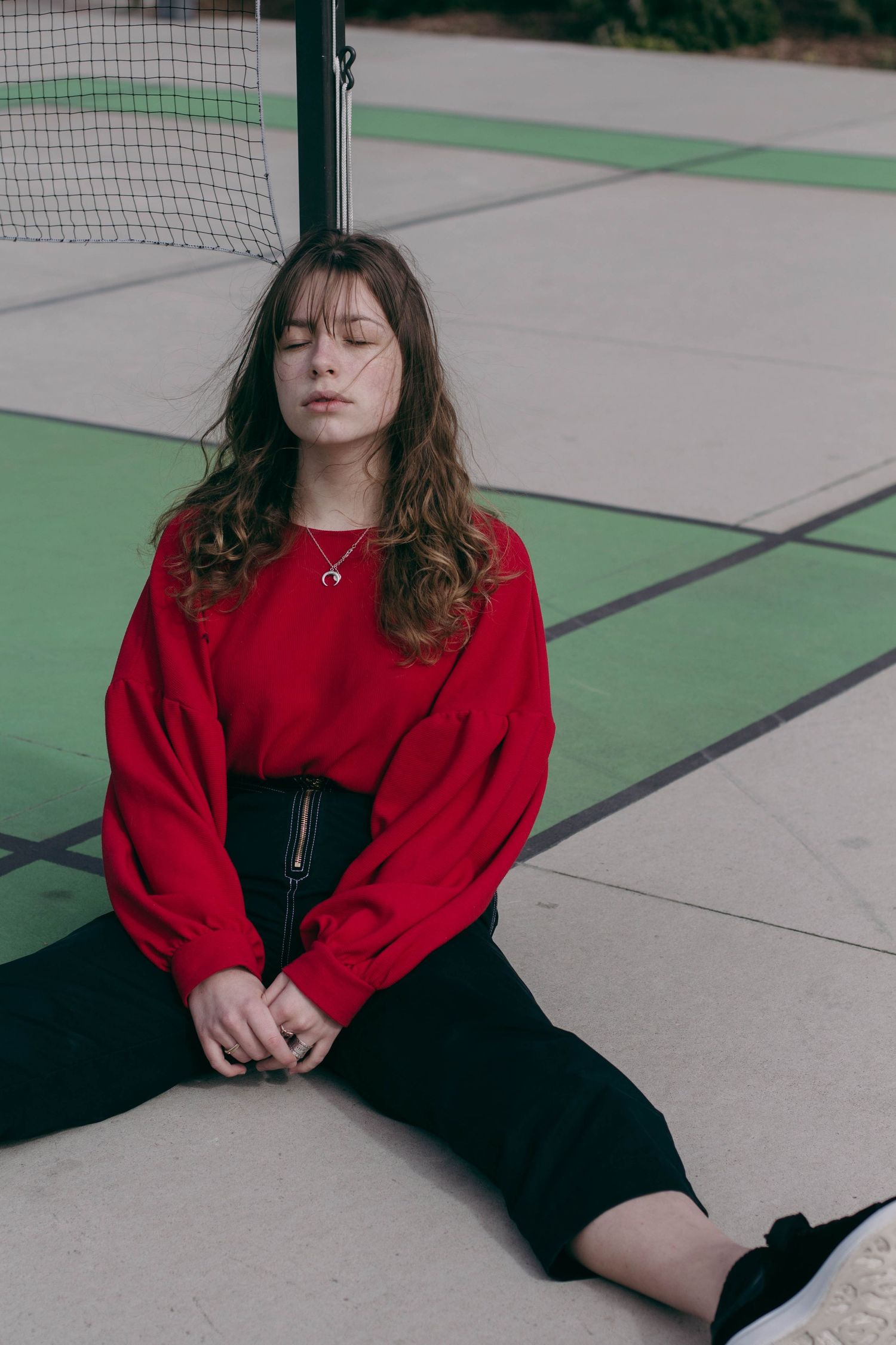 Young woman in red oversized sweater and black skirt sitting on outdoor court with eyes closed.
