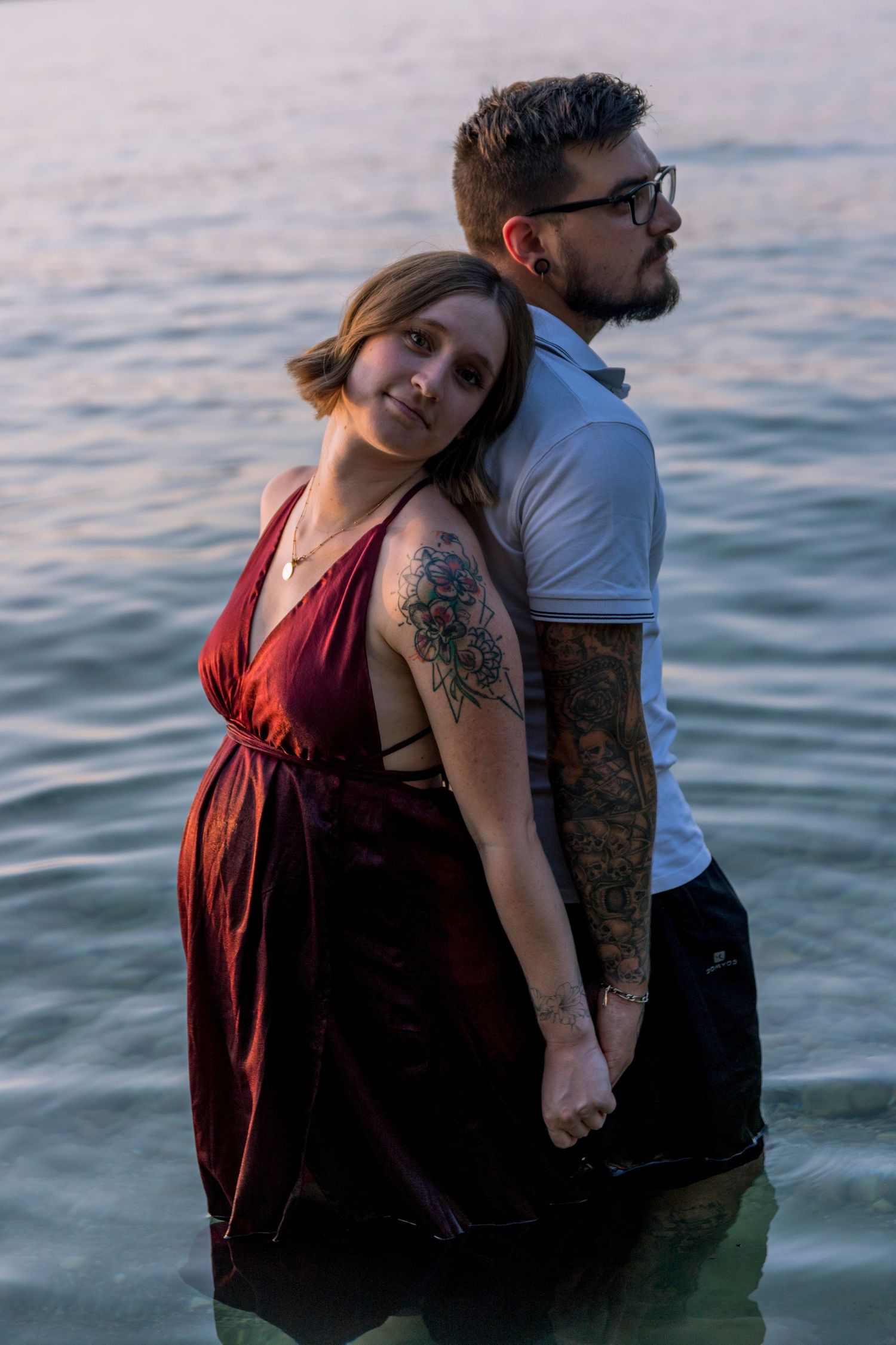Pregnant tattooed woman in red dress leaning back against tattooed partner standing in lake.
