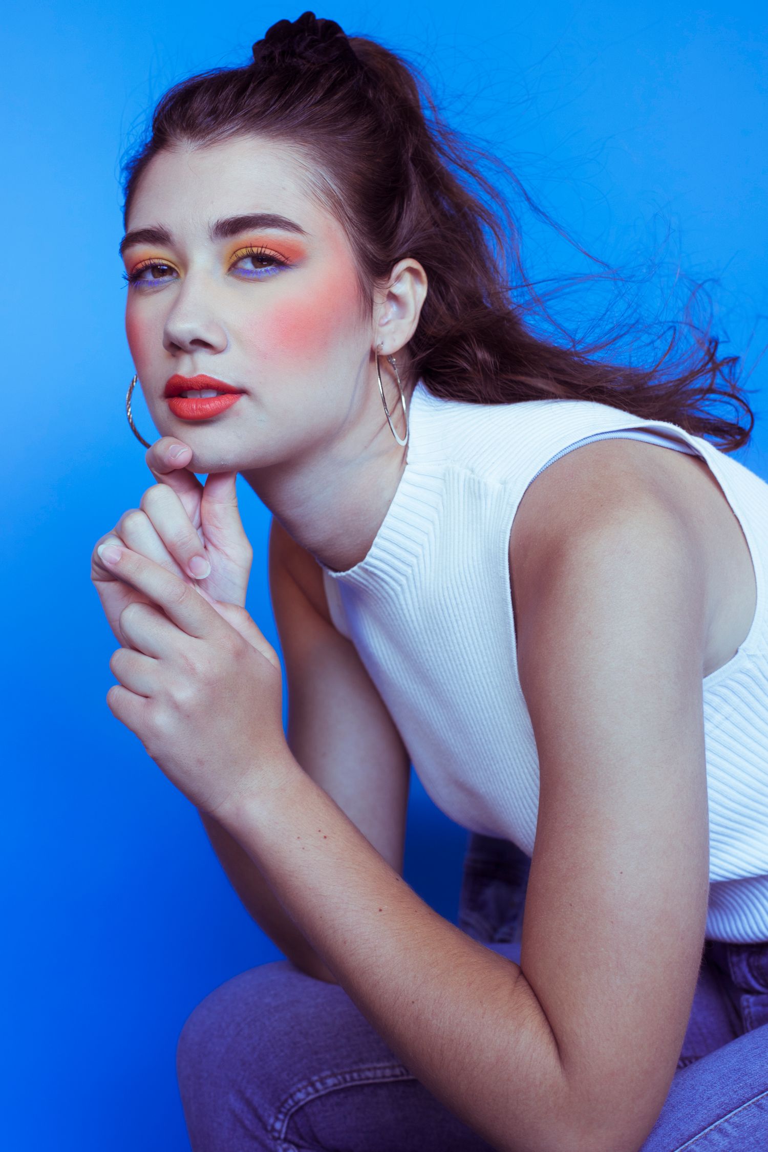 Young woman with colorful orange eye makeup poses thoughtfully with hands clasped against blue background.