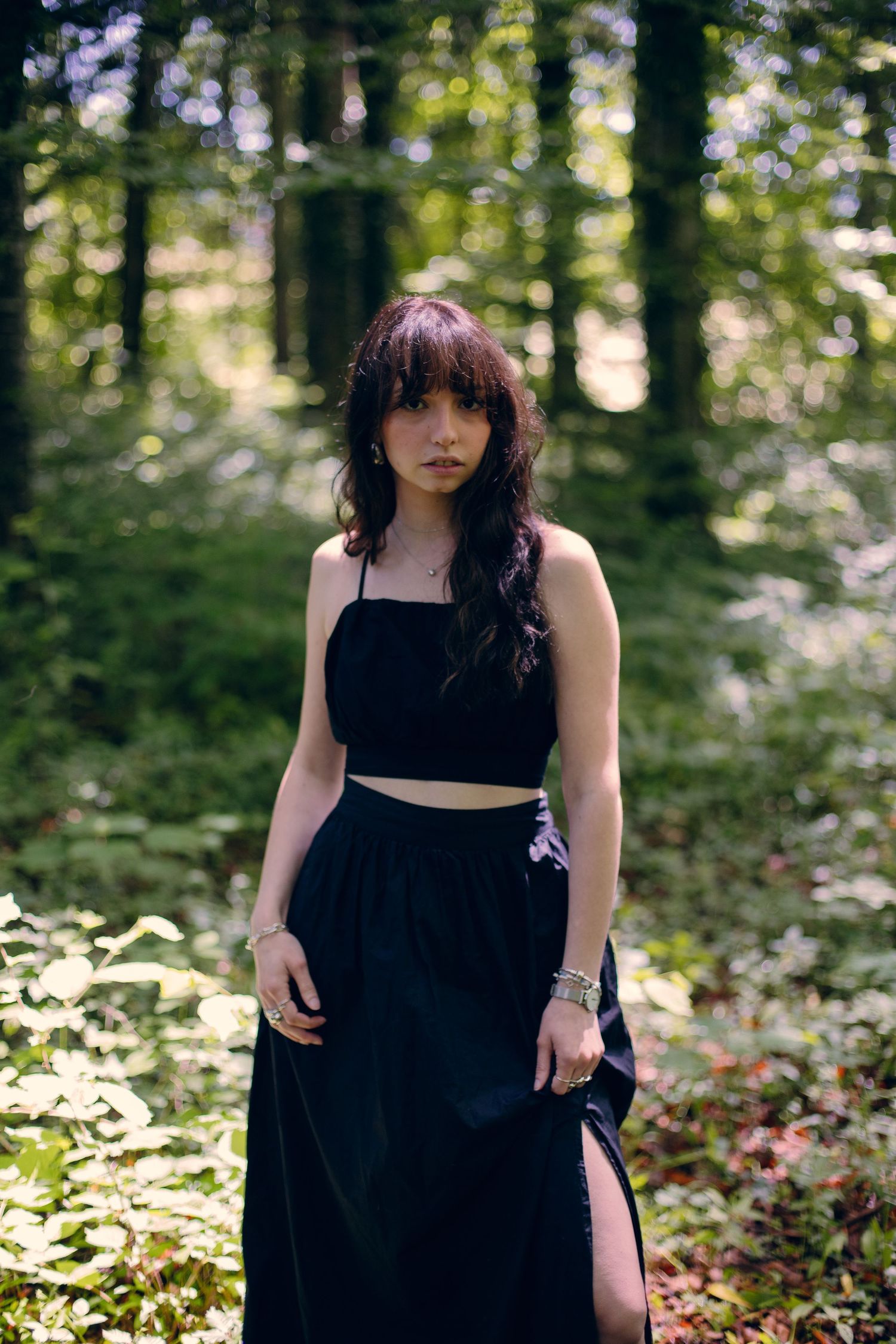 Dark-haired woman in black two-piece outfit standing among trees in forest.