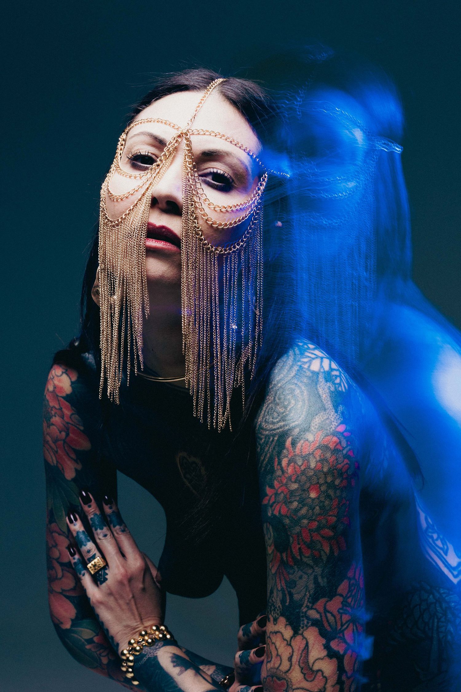 Heavily tattooed woman wearing an ornate gold chain face mask poses in blue-lit studio.
