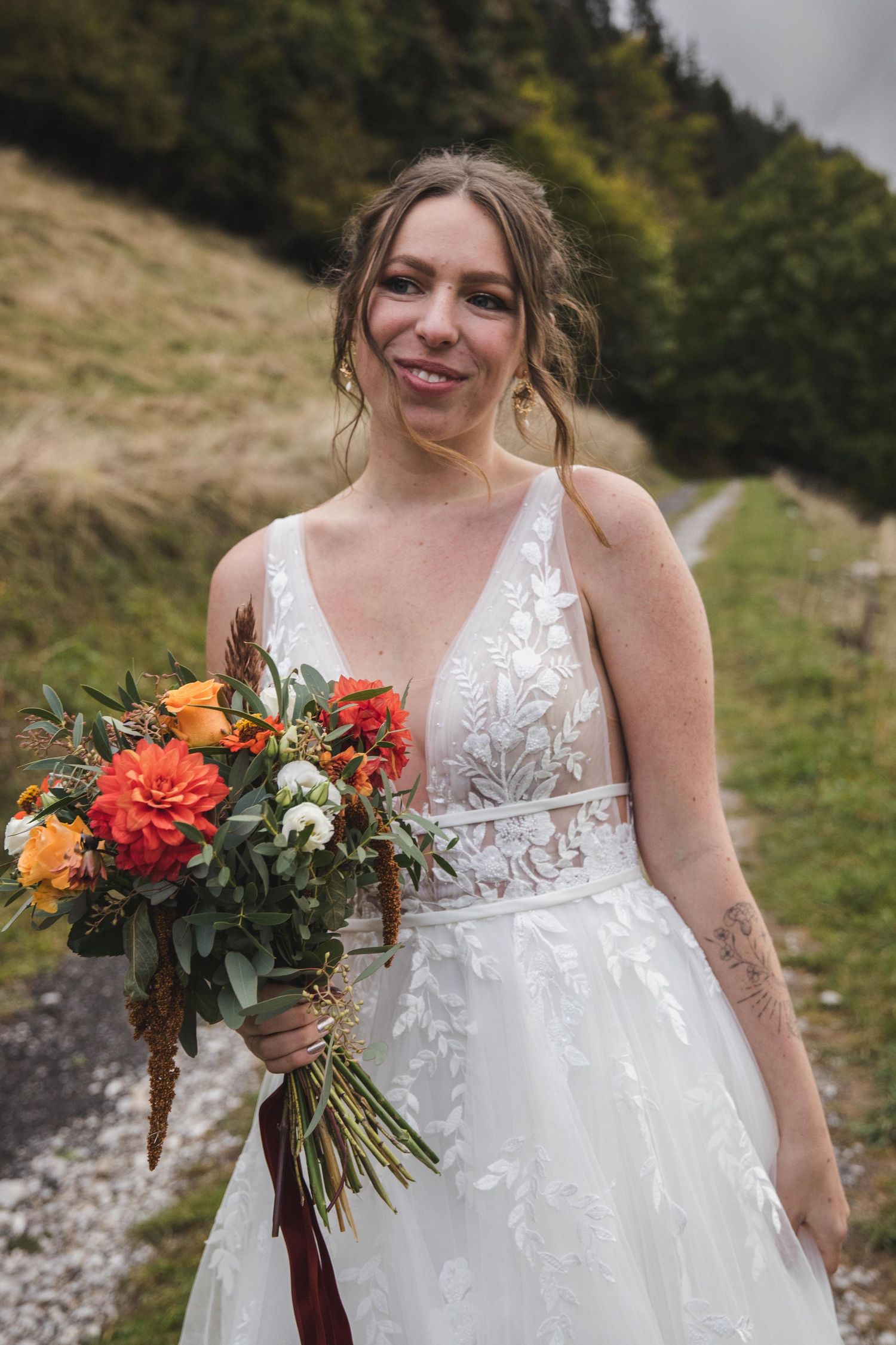 Smiling bride in lace wedding dress holding vibrant orange bouquet outdoors.