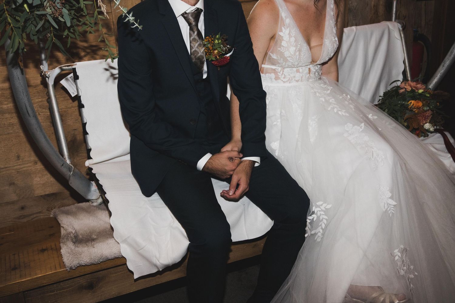 Bride in lace wedding dress and groom in dark suit sitting together at rustic barn wedding venue.