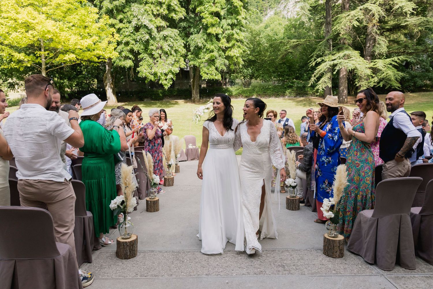 Two brides hold hands walking down the aisle at an outdoor wedding ceremony with cheering guests.