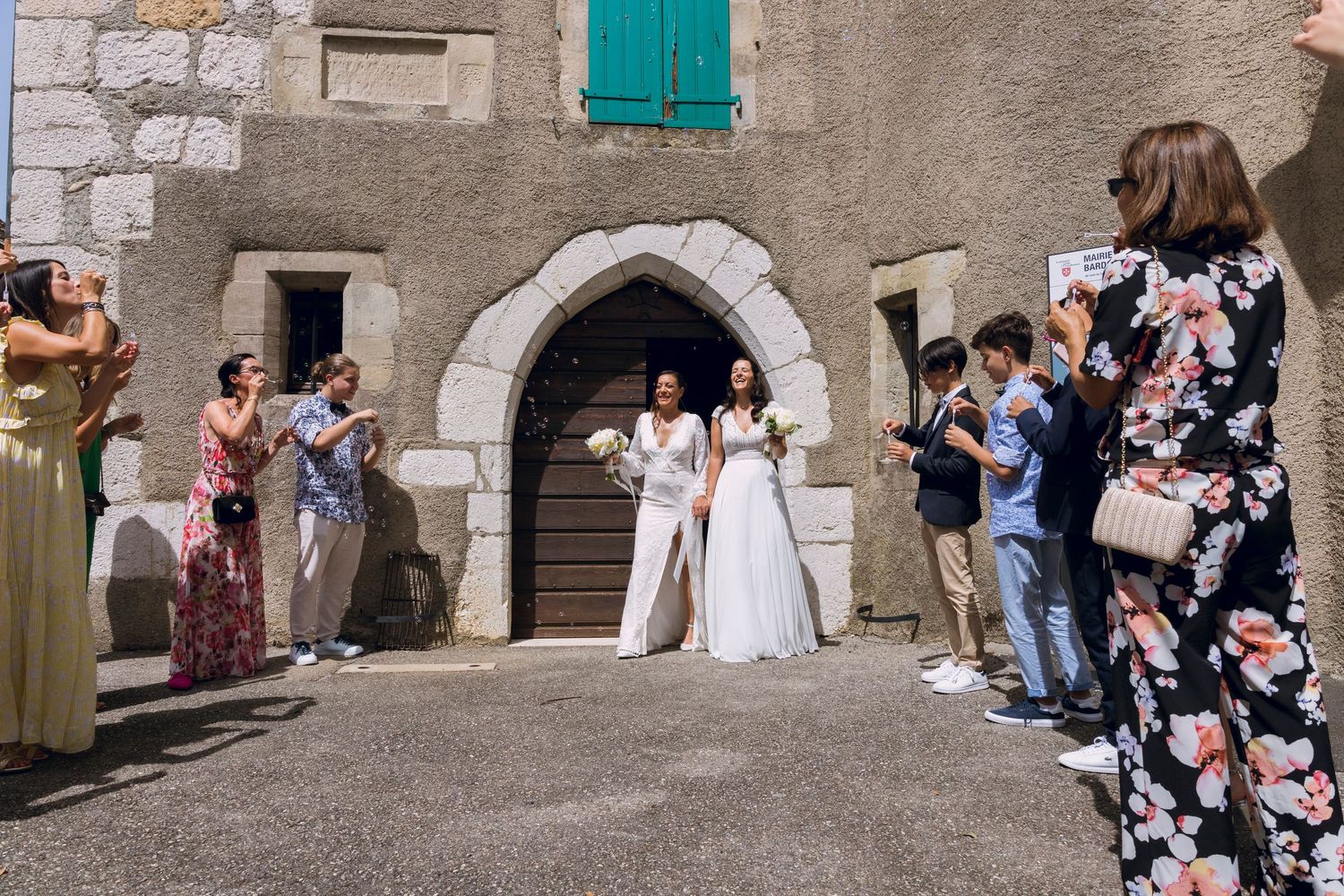 Two brides emerging from a stone archway as wedding guests celebrate and take photos outdoors.