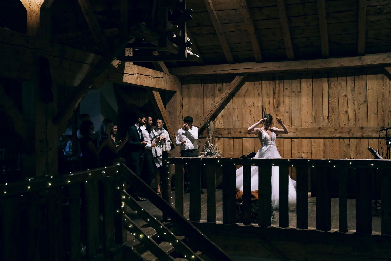 Bride in white gown stands on fairy light-adorned balcony of rustic wooden barn.