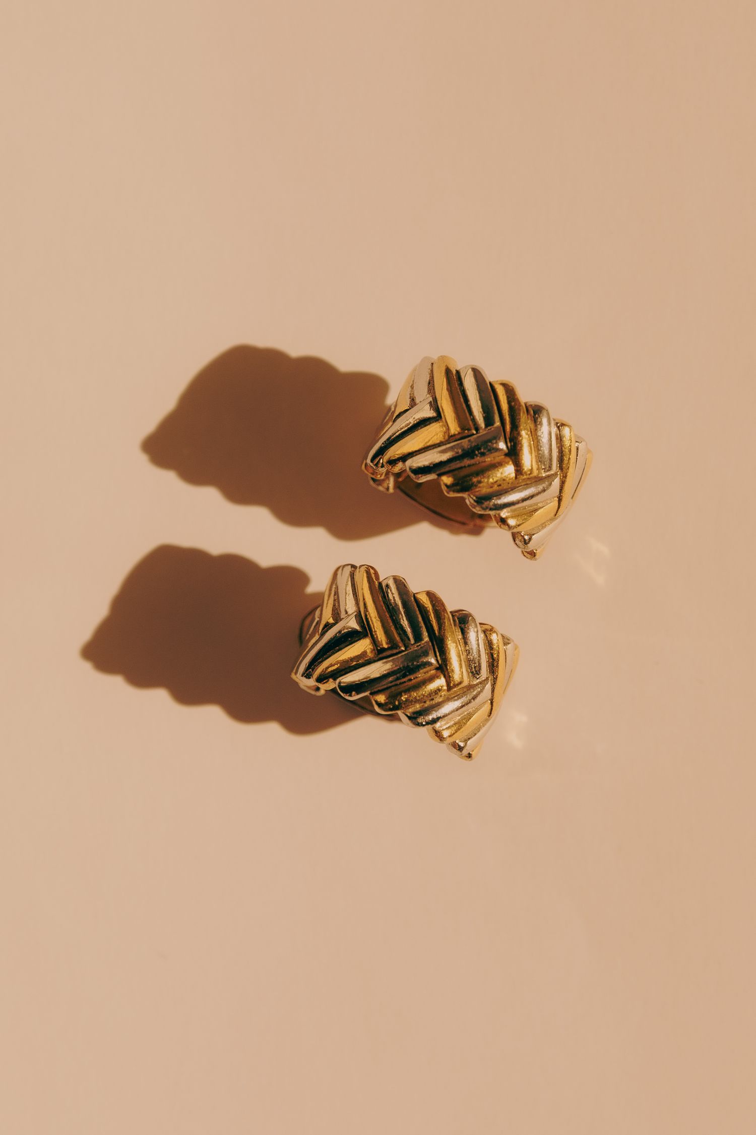 Pair of gold braided chevron clip-on earrings on a beige background with shadows.