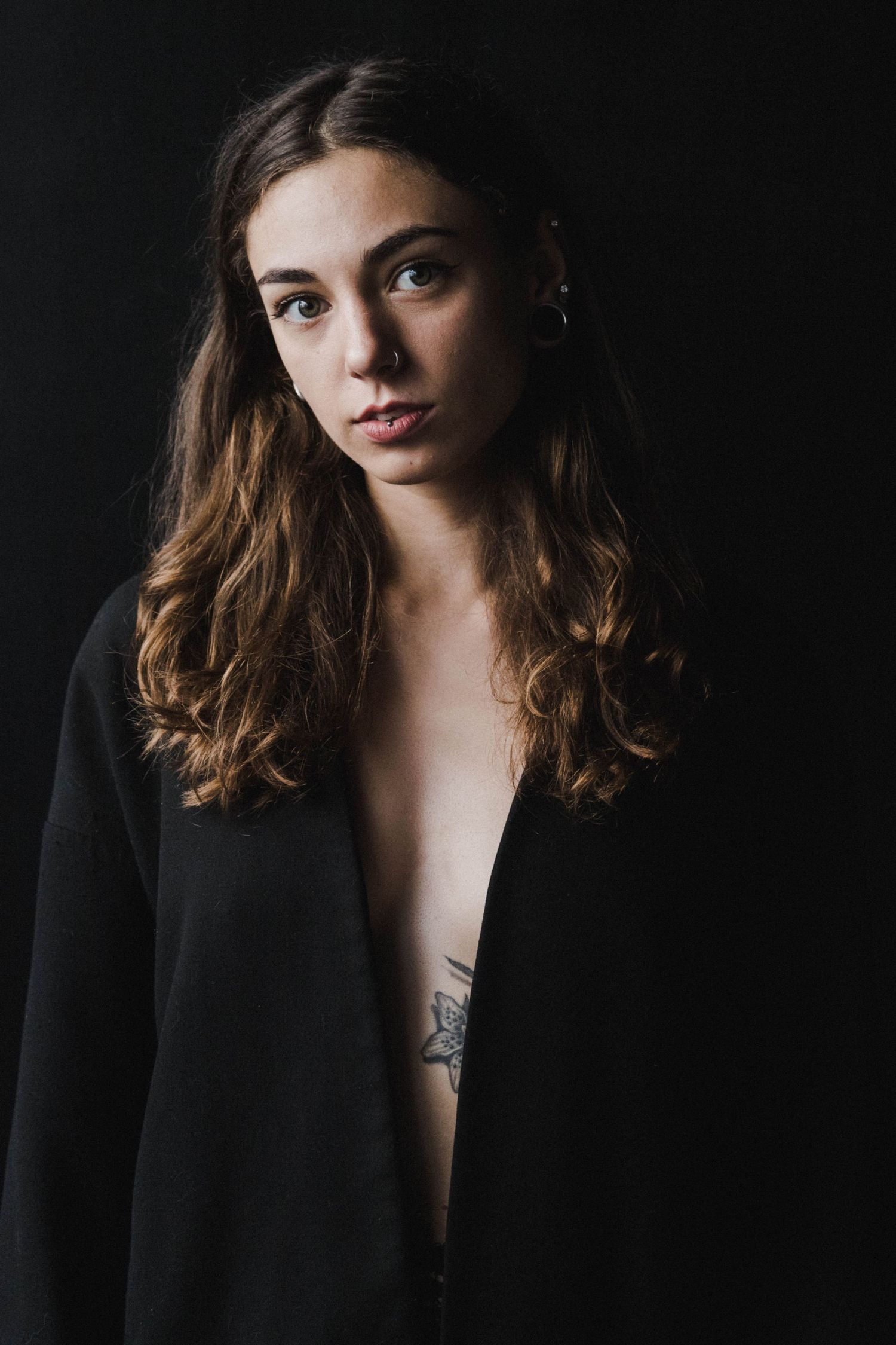 Young woman with wavy brown hair in open black cardigan revealing floral tattoo posed against black background.