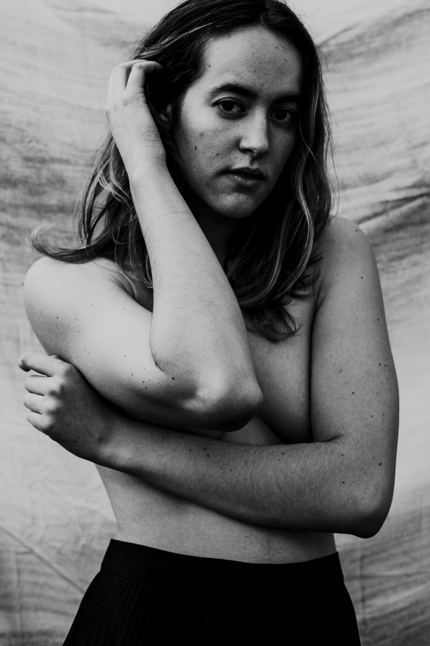 Black and white portrait of a topless woman with arms raised, hand in hair, against a draped fabric background.