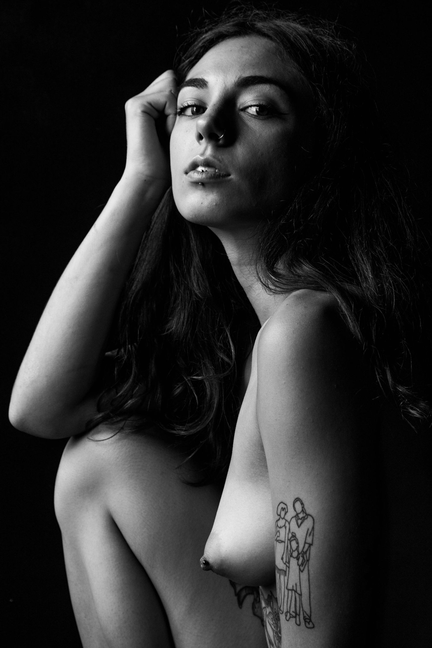 Black and white portrait of tattooed brunette woman posing nude against dark background.