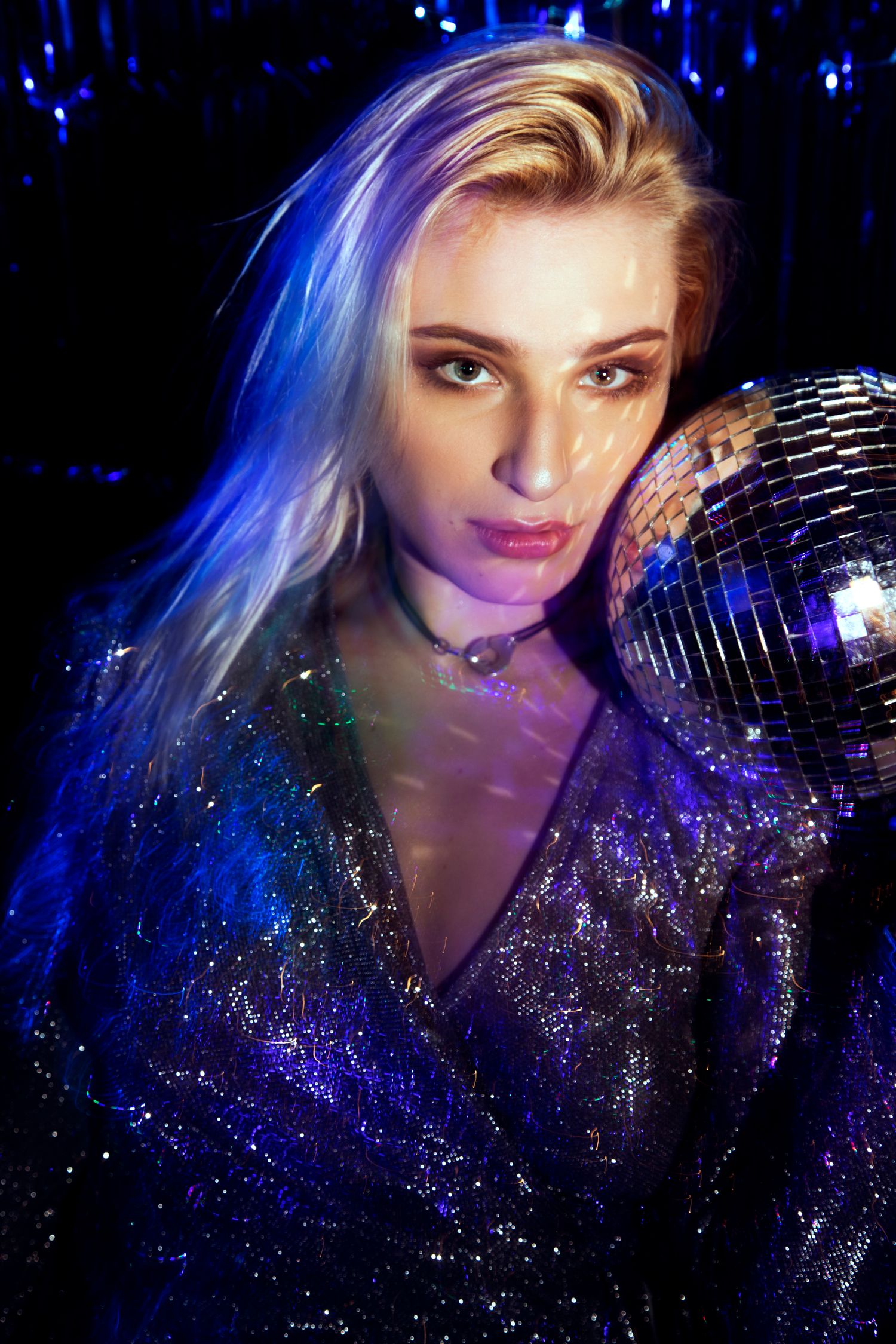 Portrait of a blonde woman in a sequined outfit leaning against a disco ball under blue club lighting.