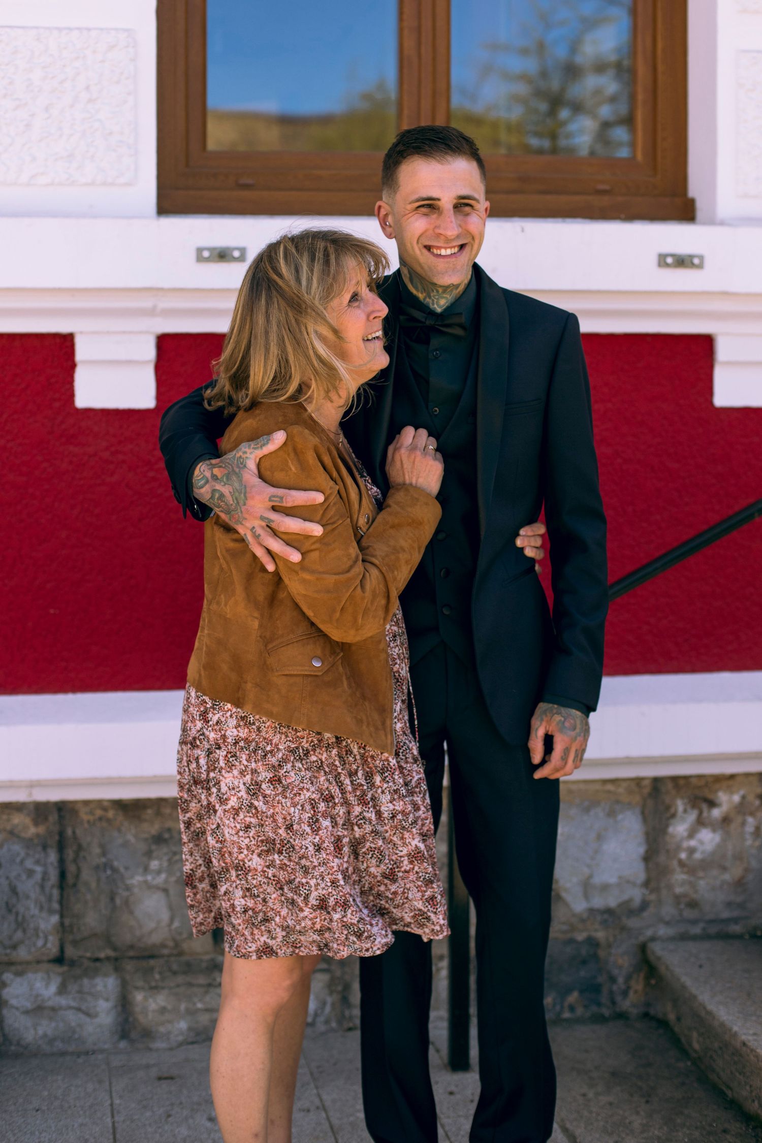 Smiling tattooed groom in black suit embraced by older blonde woman in brown jacket outside building.