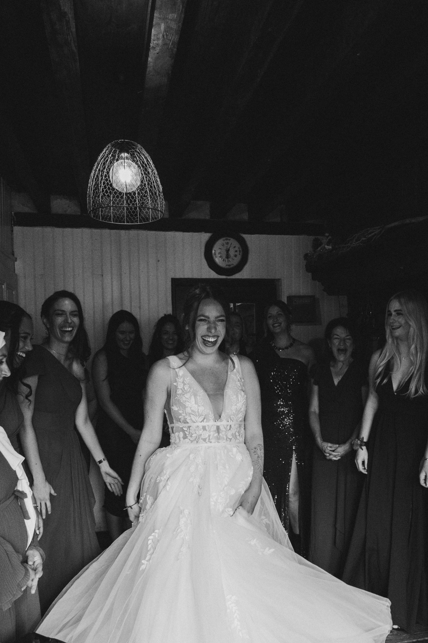 Bride laughing joyfully in a lace wedding gown surrounded by smiling guests at an indoor wedding reception.