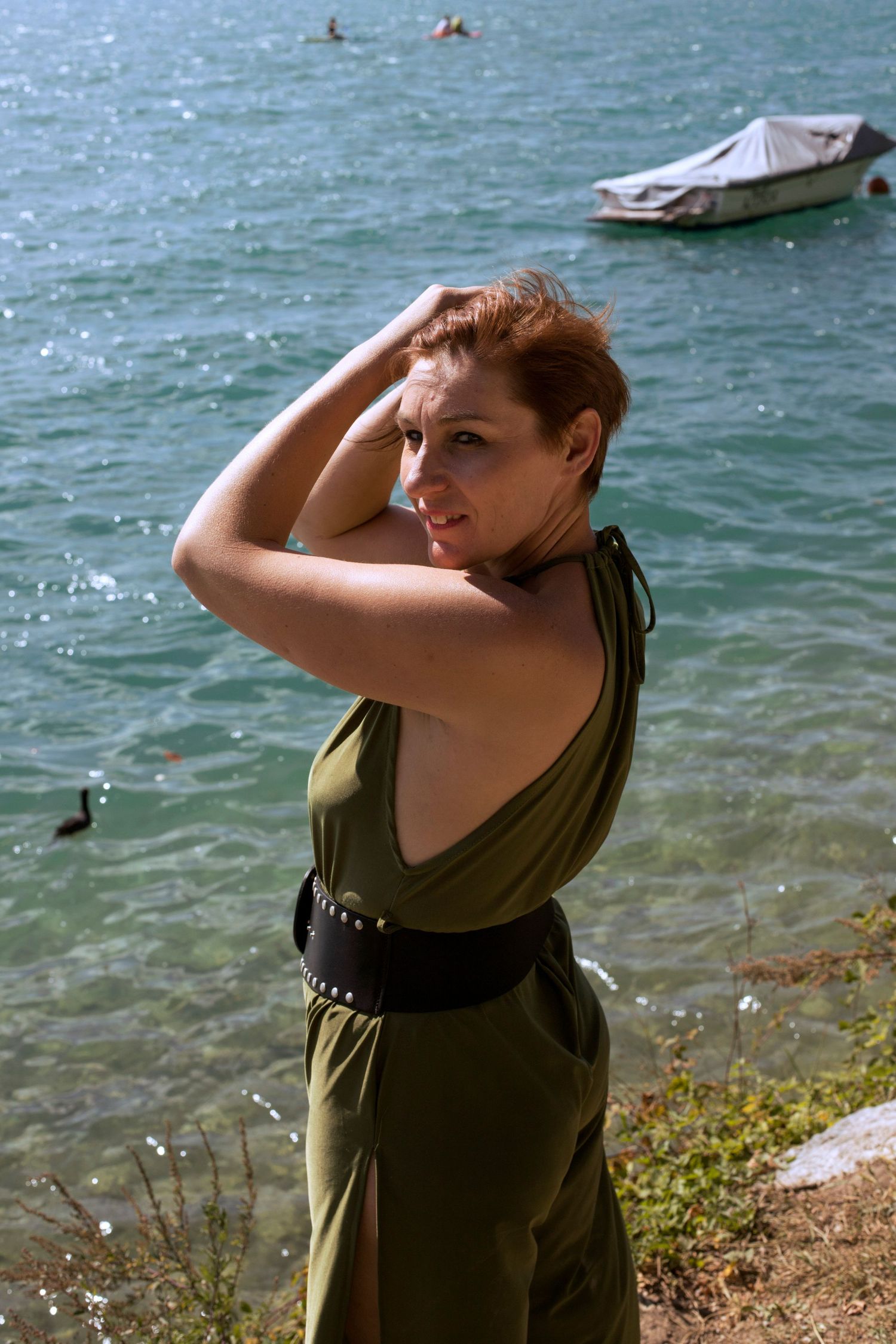 Woman in olive green halter dress with black belt posing by a scenic turquoise lake with a boat.