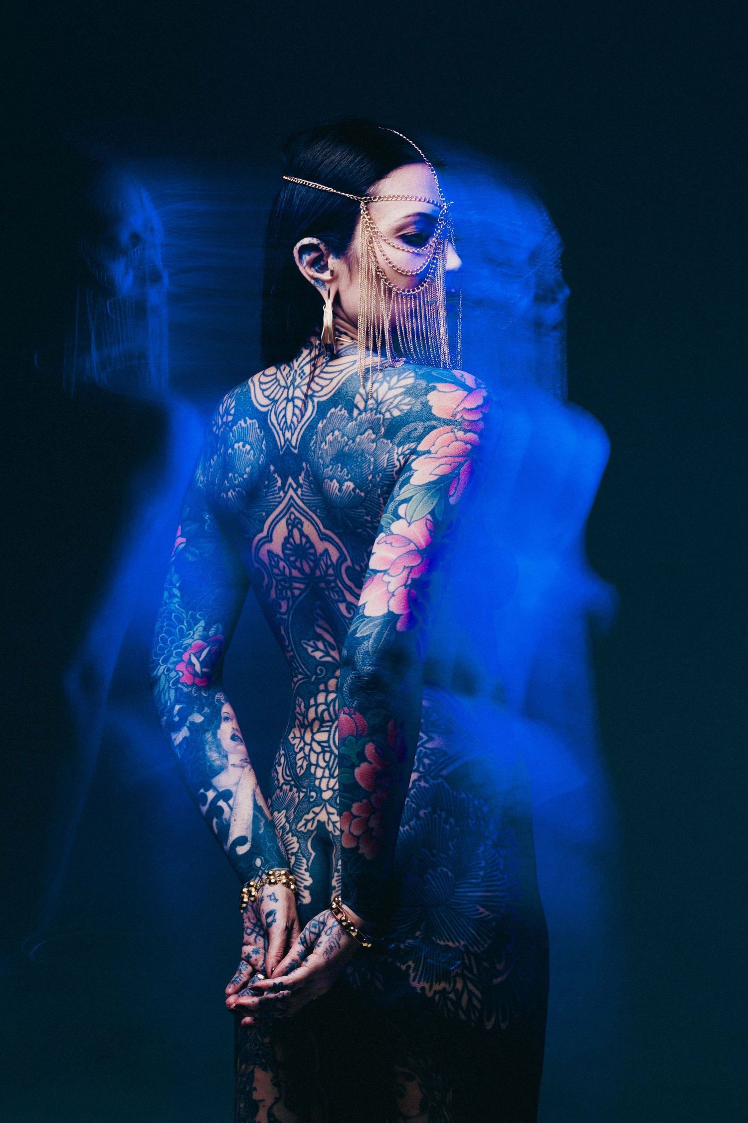 Tattooed woman in profile wearing gold chain headpiece with blue motion blur effect in studio.