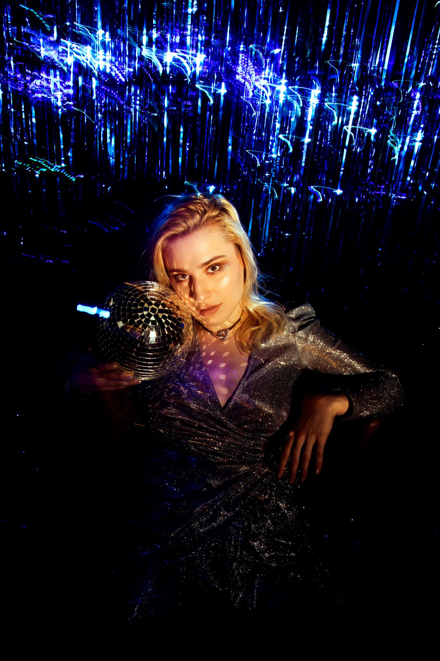 Blonde woman in glittery silver dress poses with disco ball against blue neon light streaks.