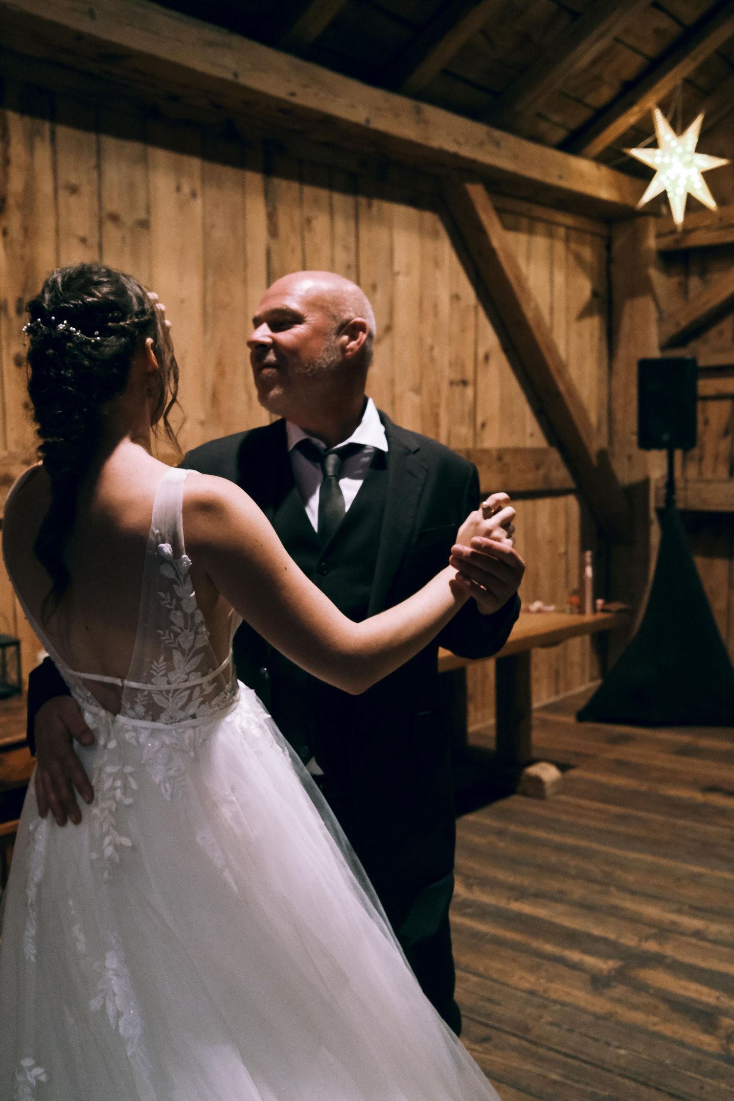 Bride in white lace gown shares first dance with suited partner in warmly lit wooden barn venue.