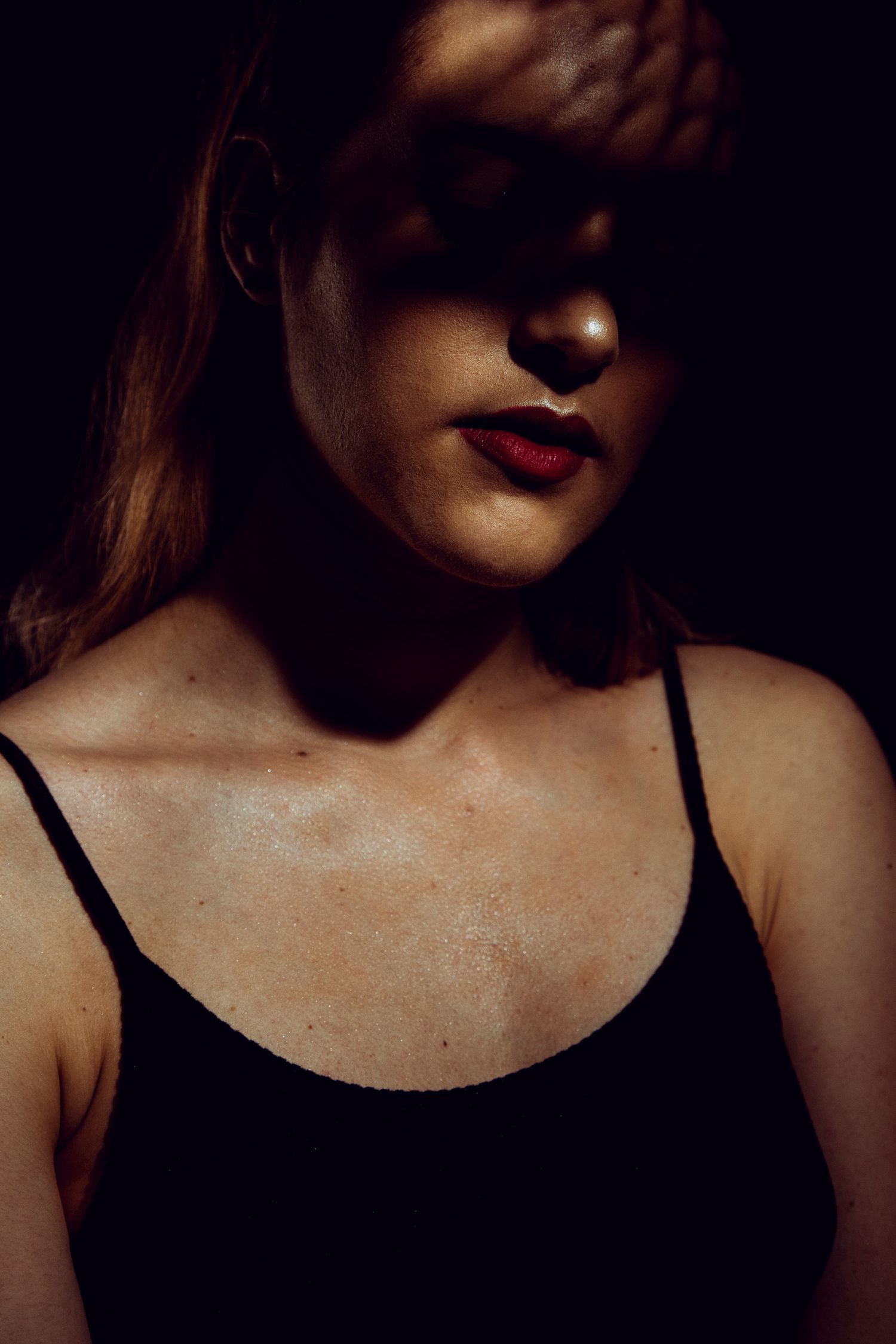 Dark artistic portrait of a woman with red lips wearing black tank top with shadow obscuring her eyes.