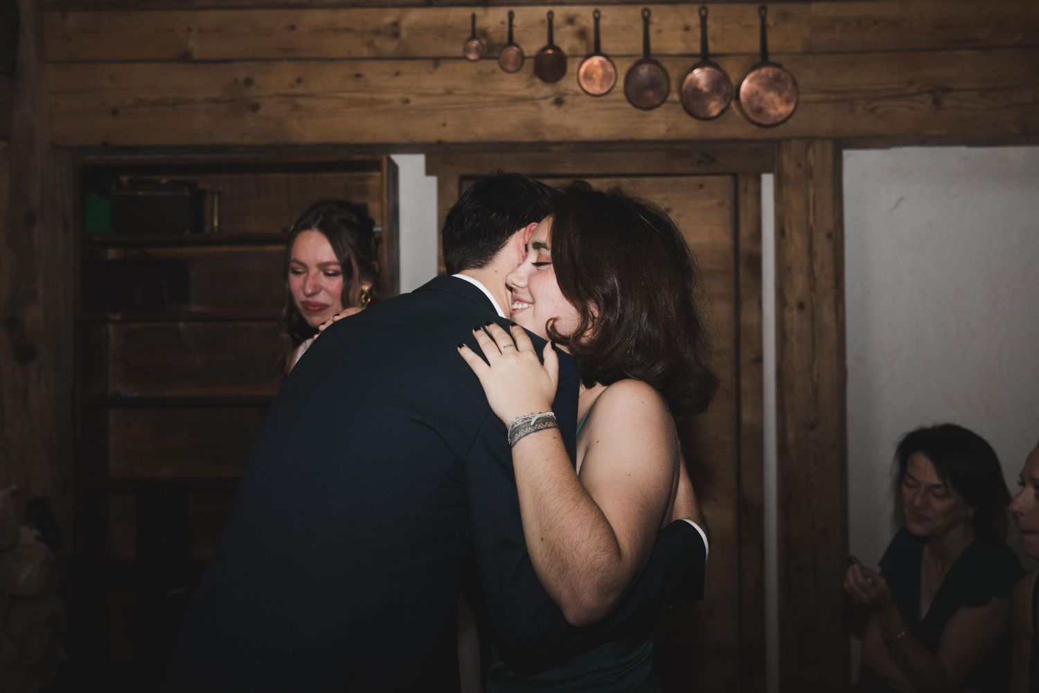 Couple sharing a romantic kiss at wedding reception while guests celebrate in rustic barn setting.