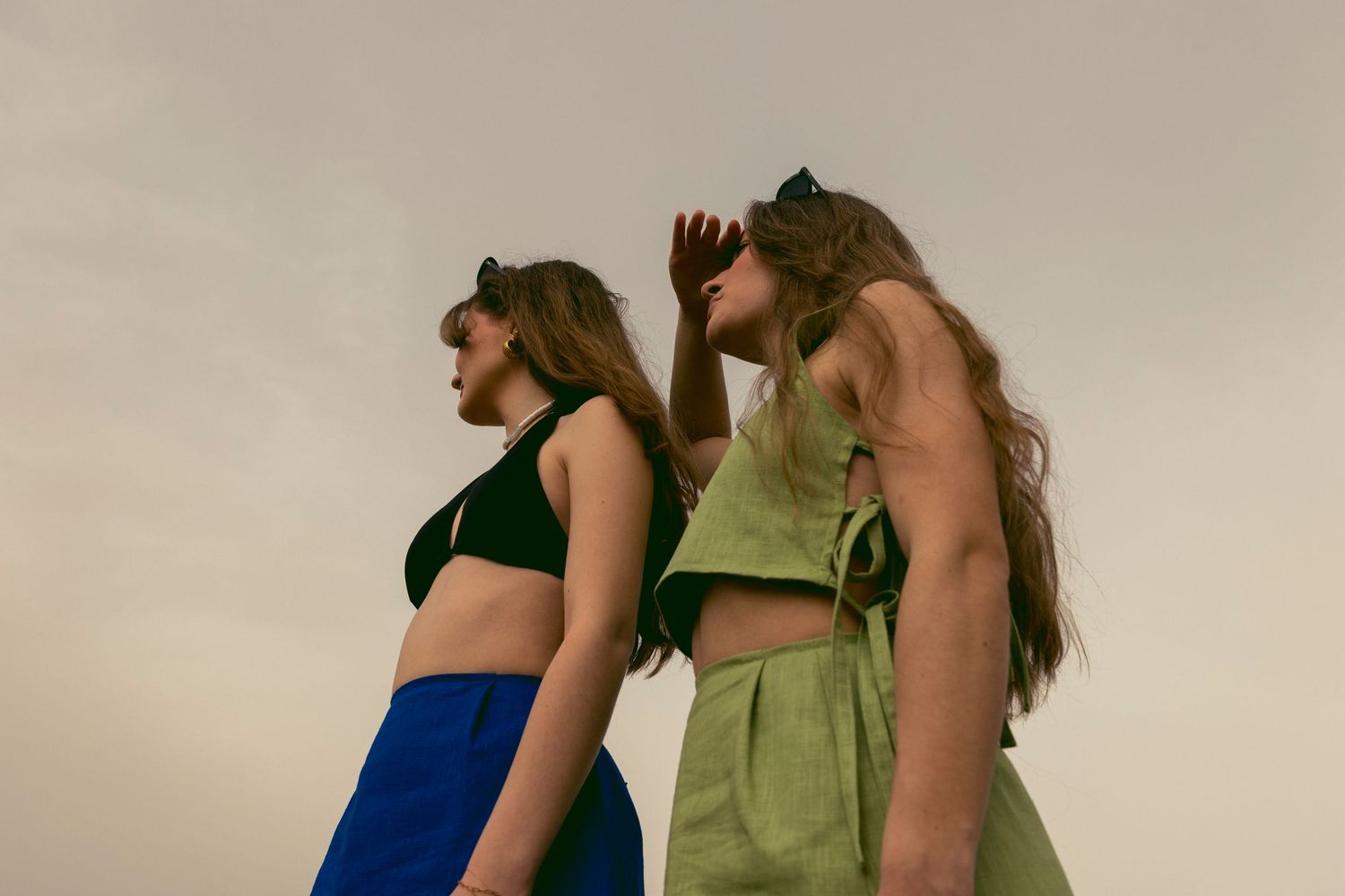 Two young women in colorful crop tops and skirts pose outdoors against a pale overcast sky.