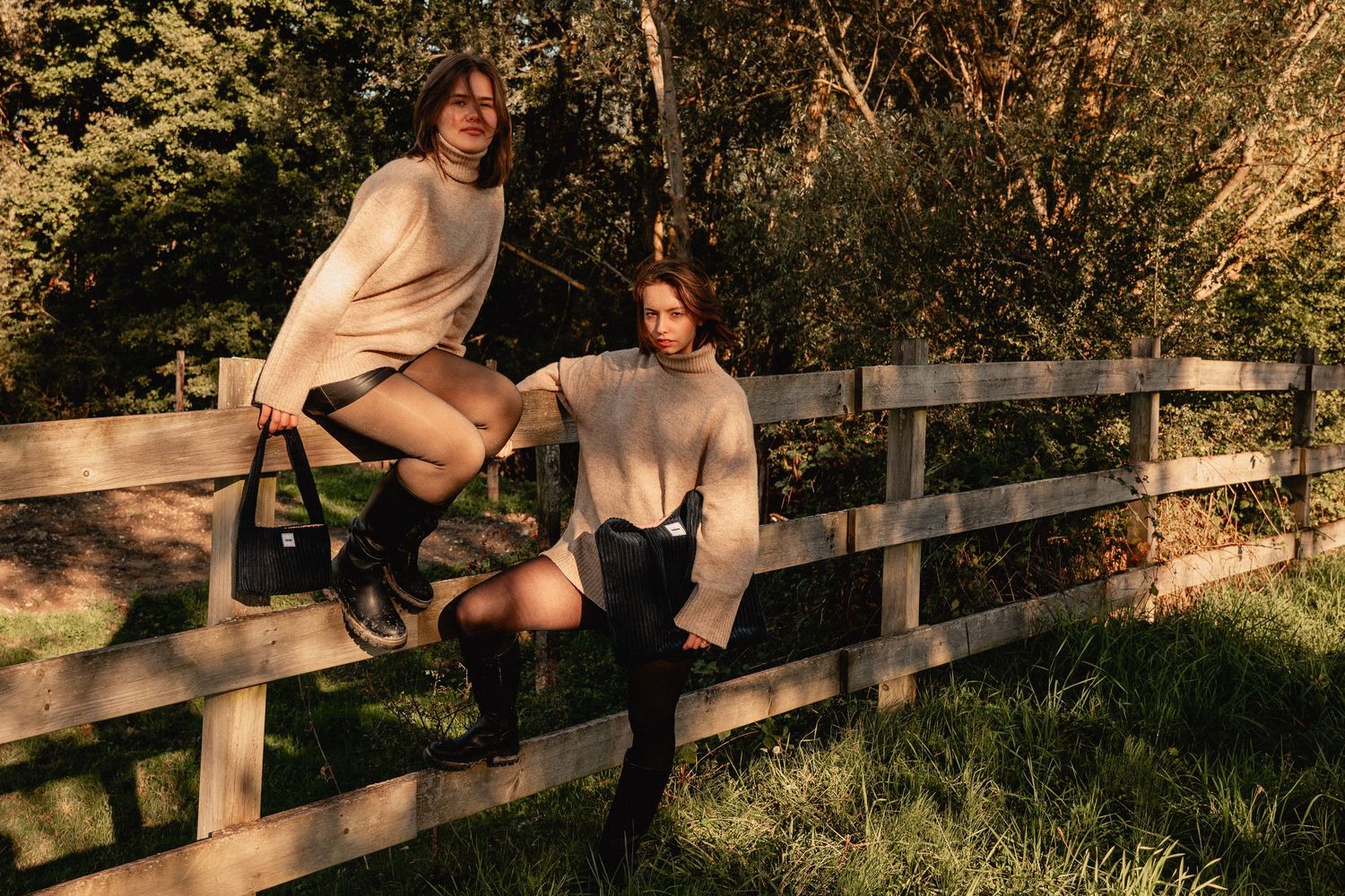 Two women in cream oversized sweaters sit and lean on a rustic wooden fence in a lush outdoor setting.