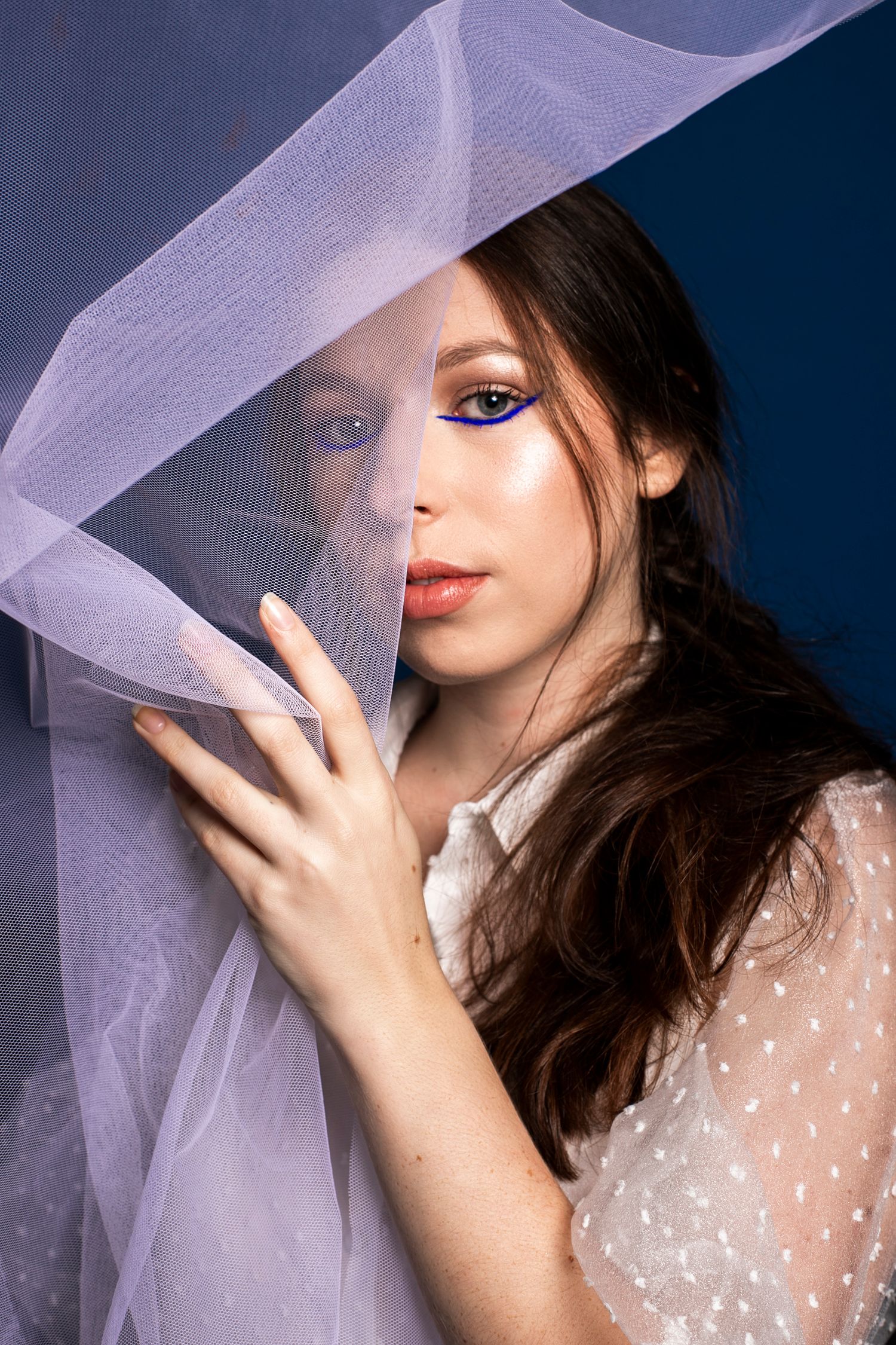 Brunette woman with colorful eye makeup holding a white sheer veil against a blue background.