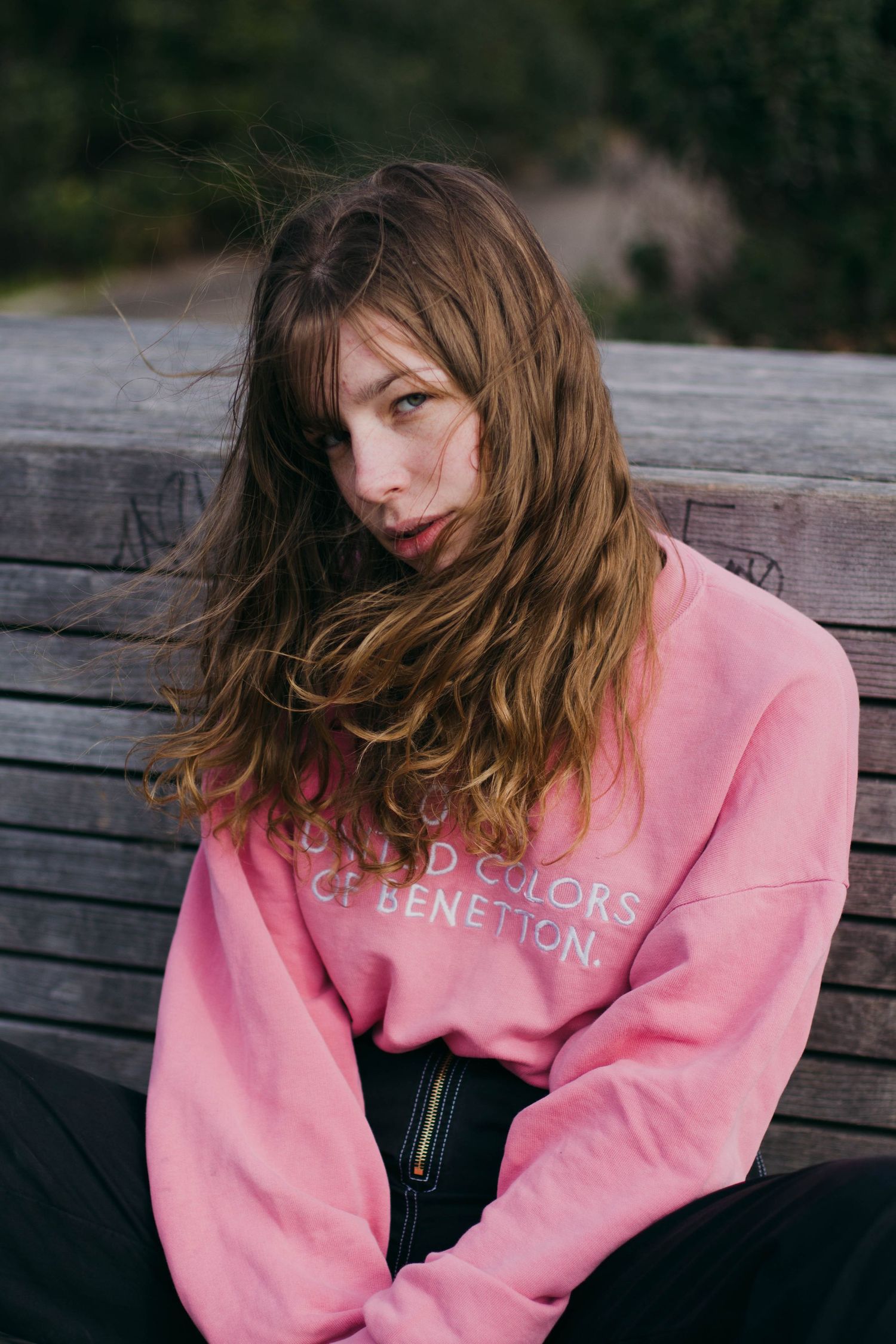 Young woman with windswept hair wearing pink United Colors of Benetton sweatshirt sitting on wooden bench outdoors.