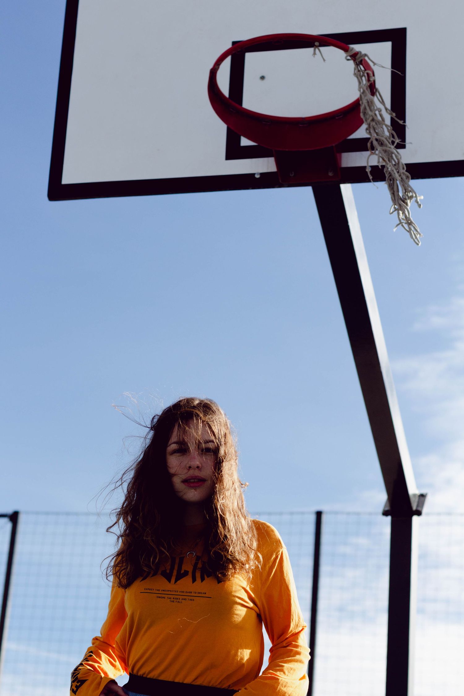 Young woman in orange long-sleeve shirt standing under basketball hoop on outdoor court against blue sky.