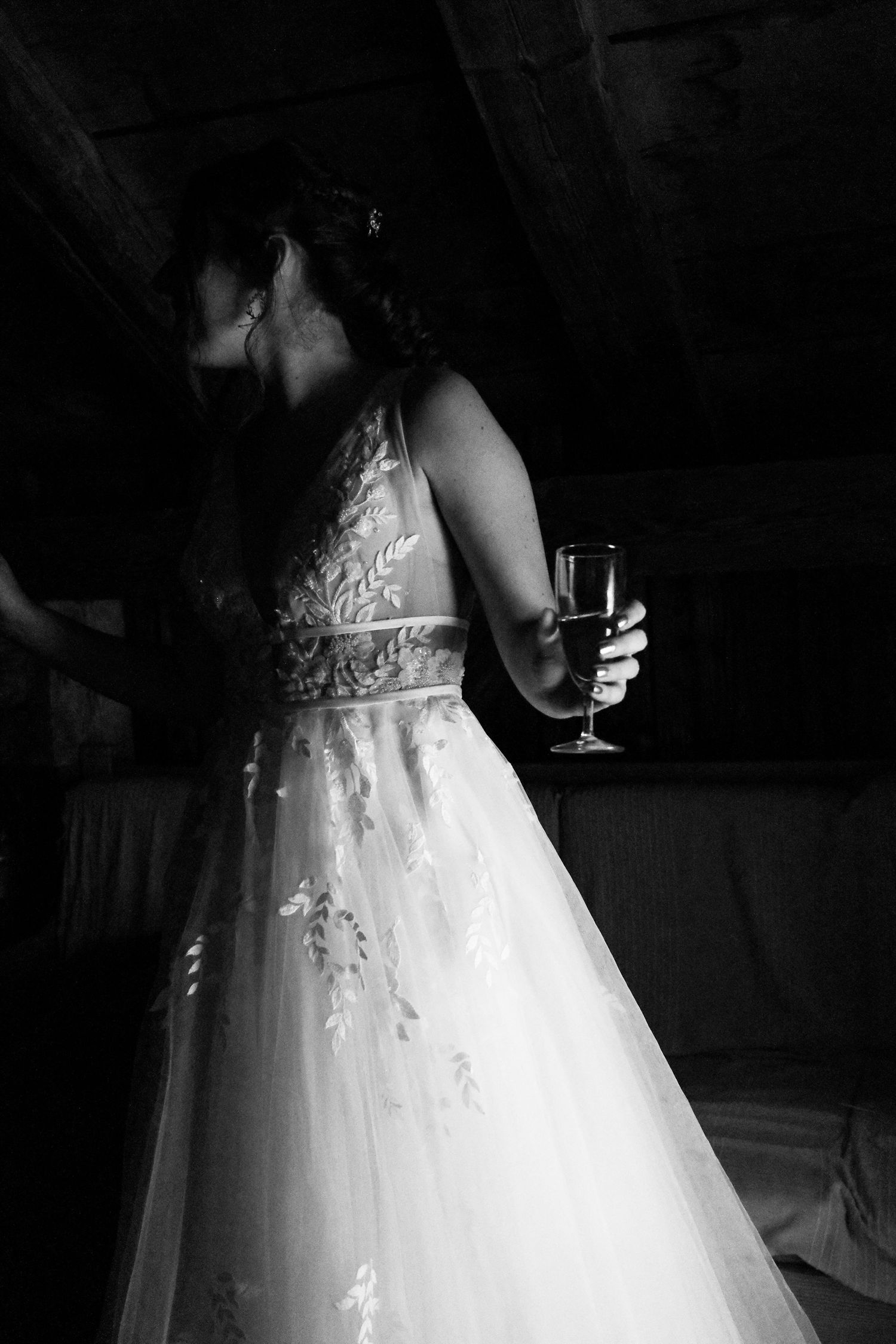 Black and white portrait of bride in embroidered gown holding champagne glass in dimly lit barn.