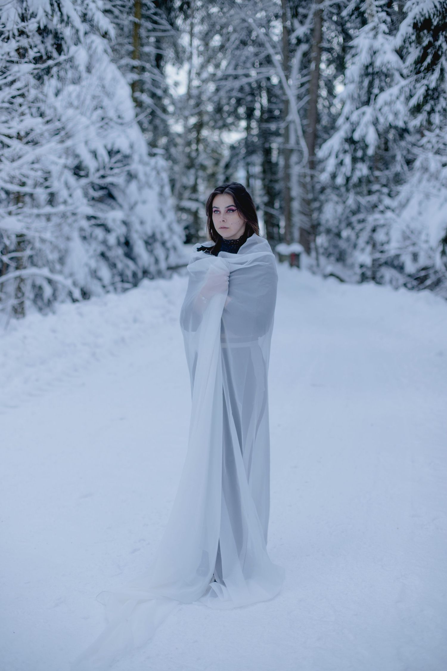 Young woman wrapped in flowing white fabric standing in a snowy winter forest path.