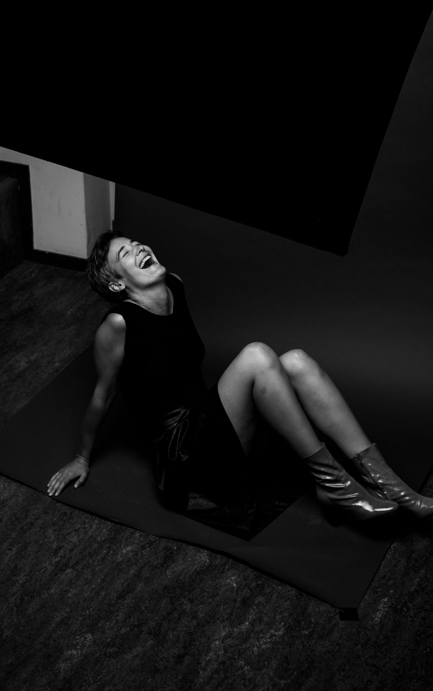 Black and white photo of laughing woman in black dress lying on floor against geometric dark backdrop.