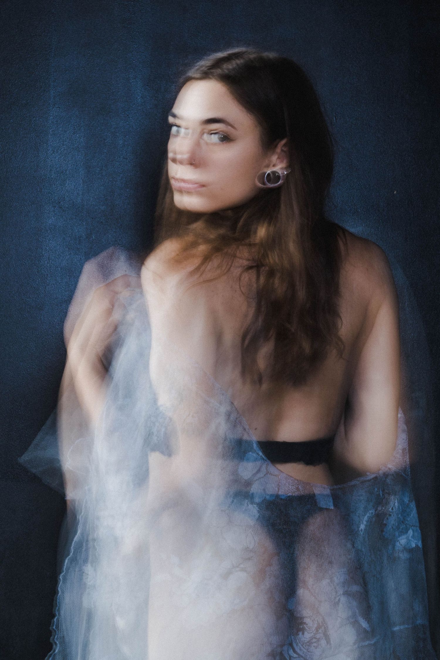 Soft focus color portrait of woman in black lingerie with sheer fabric overlay on blue background.