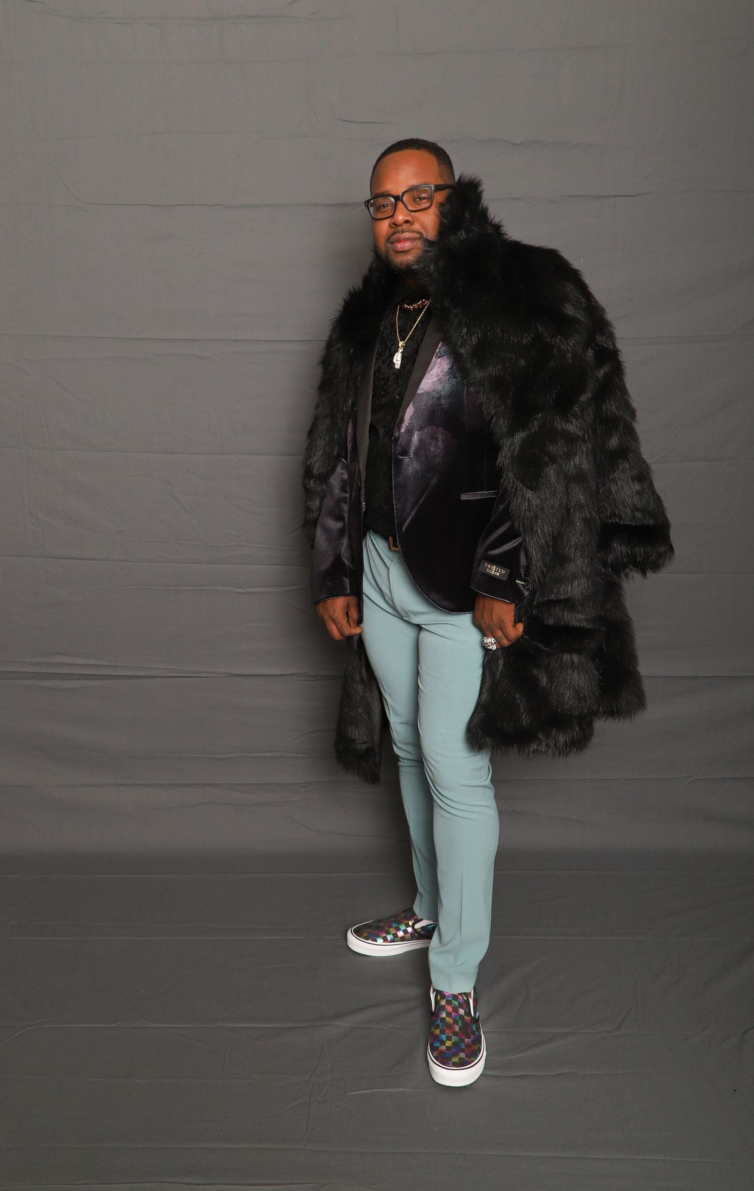 Fashionable portrait of man wearing black faux fur coat, mint green pants, and checkered sneakers against gray backdrop.