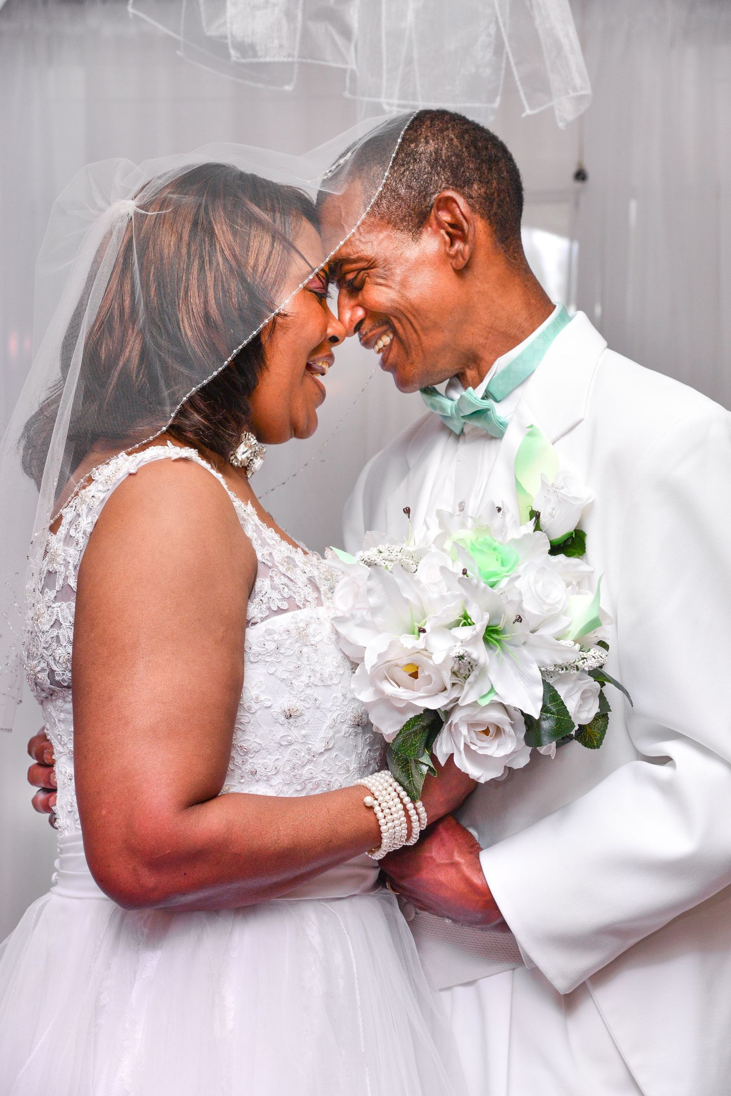 A joyful bride and groom touch foreheads romantically under a wedding veil with white roses.