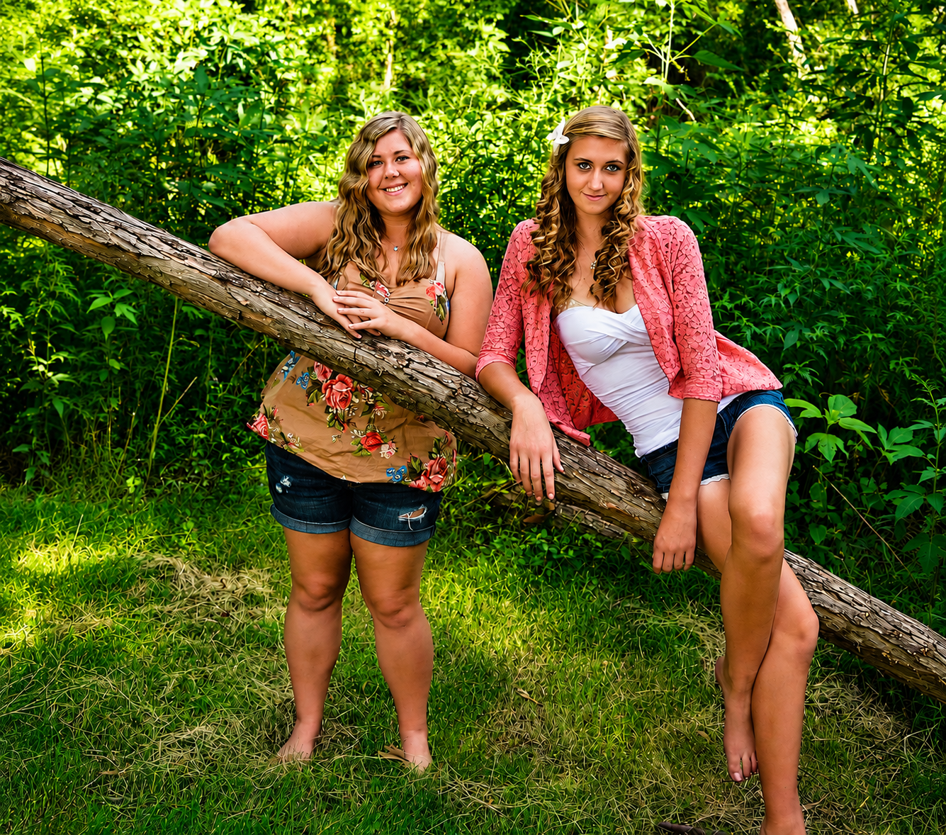 Two teenage girls posing outdoors leaning on fallen tree log surrounded by lush green foliage.