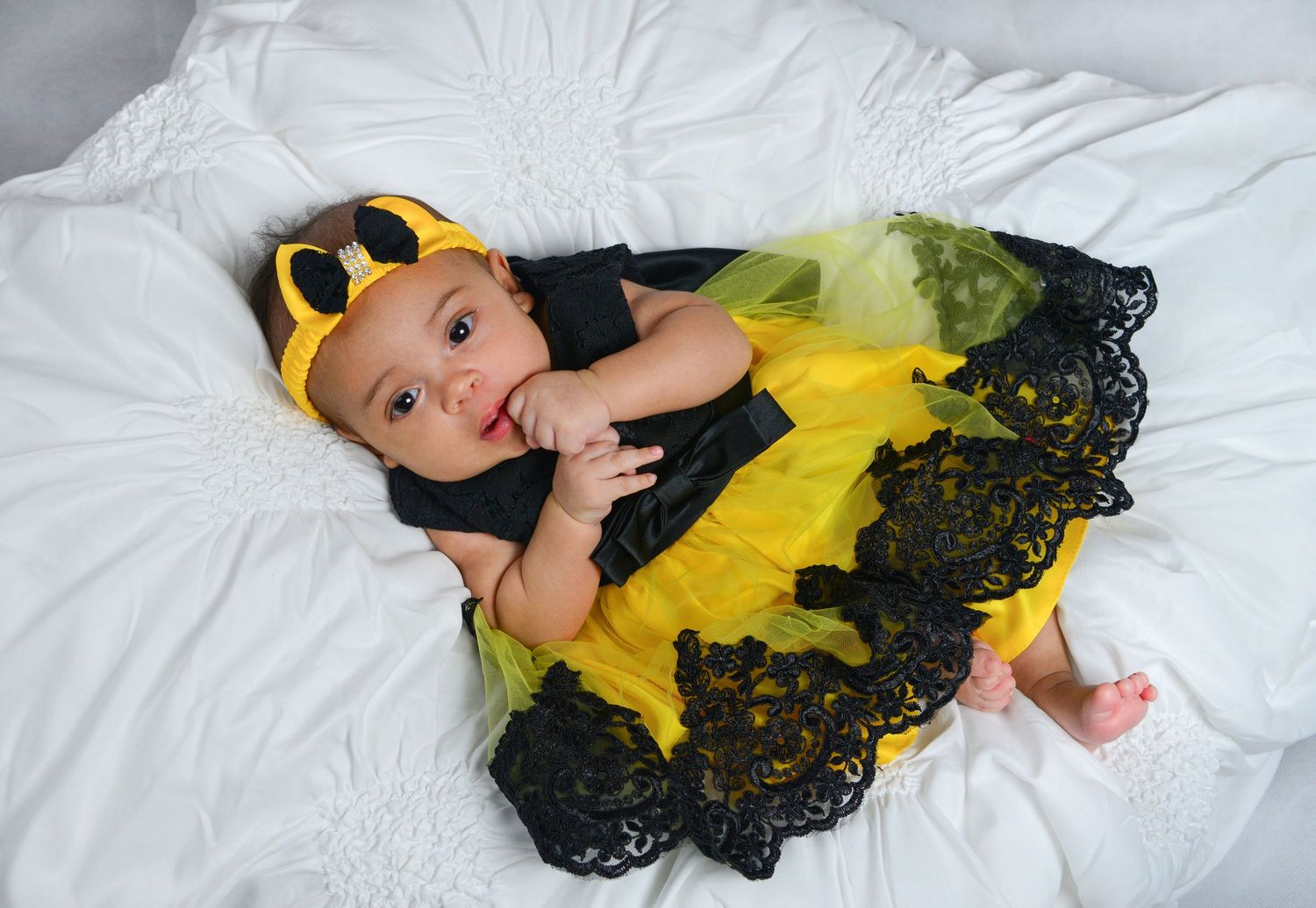 Adorable baby in a yellow and black bumblebee costume with lace trim lying on white bedding.