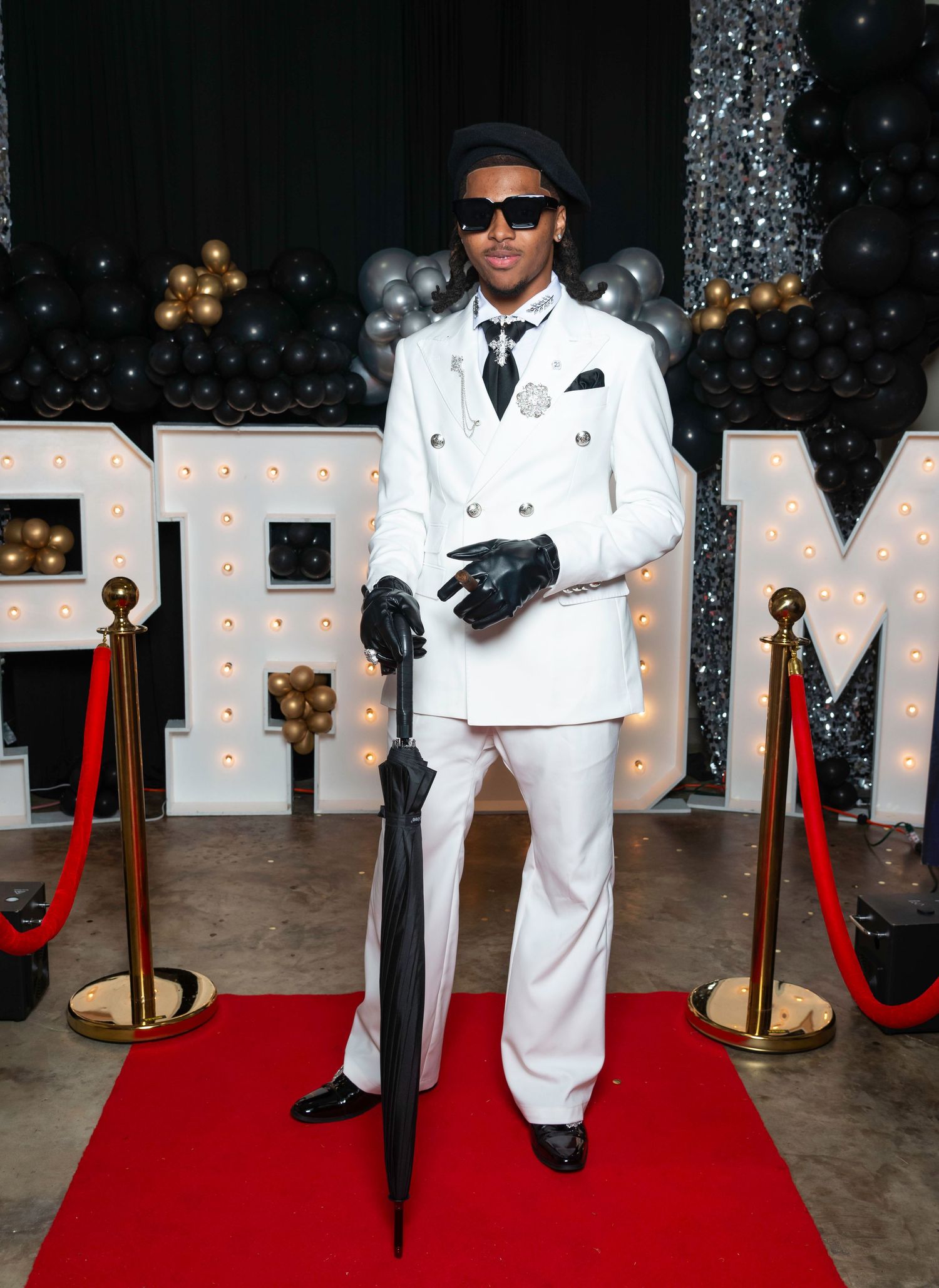 Dapper man in white suit, black hat, and gloves holds umbrella on red carpet at prom venue.