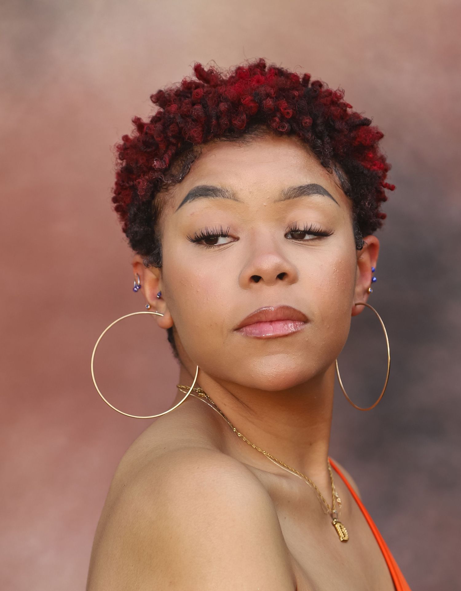 Young woman with red dyed natural curly hair wearing large gold hoop earrings and orange top.