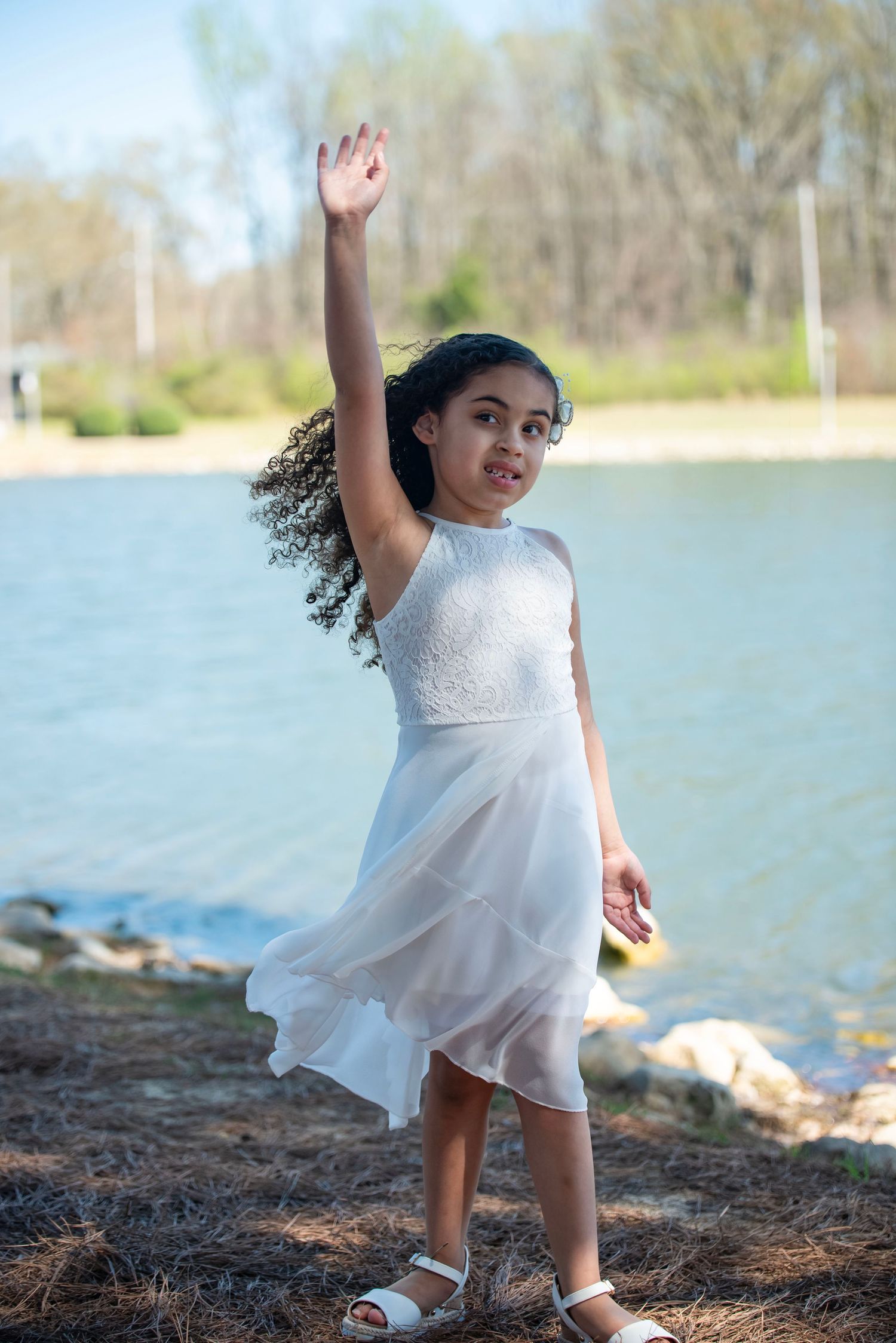 Young girl in white dress dancing by a lake with one arm raised in the air.
