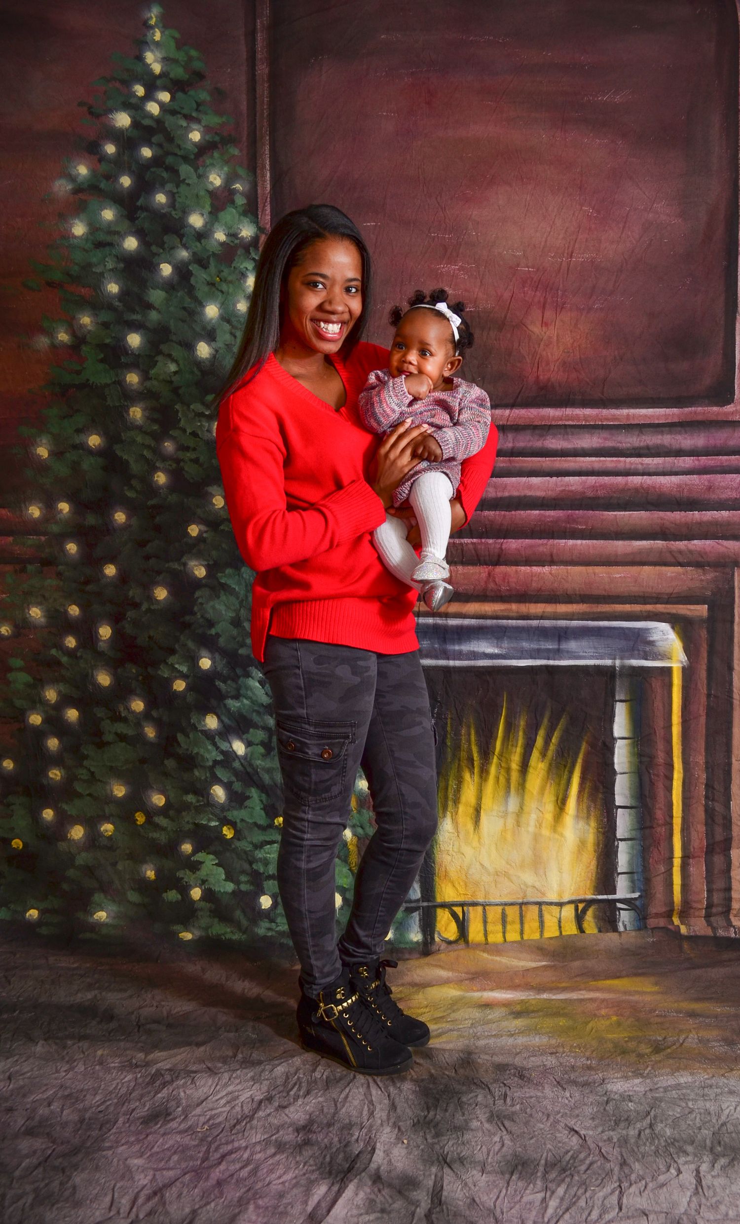 Smiling woman in red sweater holding baby girl in front of Christmas tree backdrop.