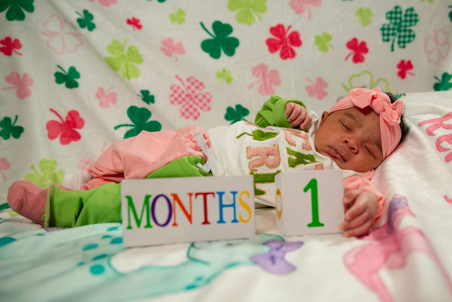 Sleeping newborn baby in pink outfit beside month milestone blocks on a shamrock blanket.