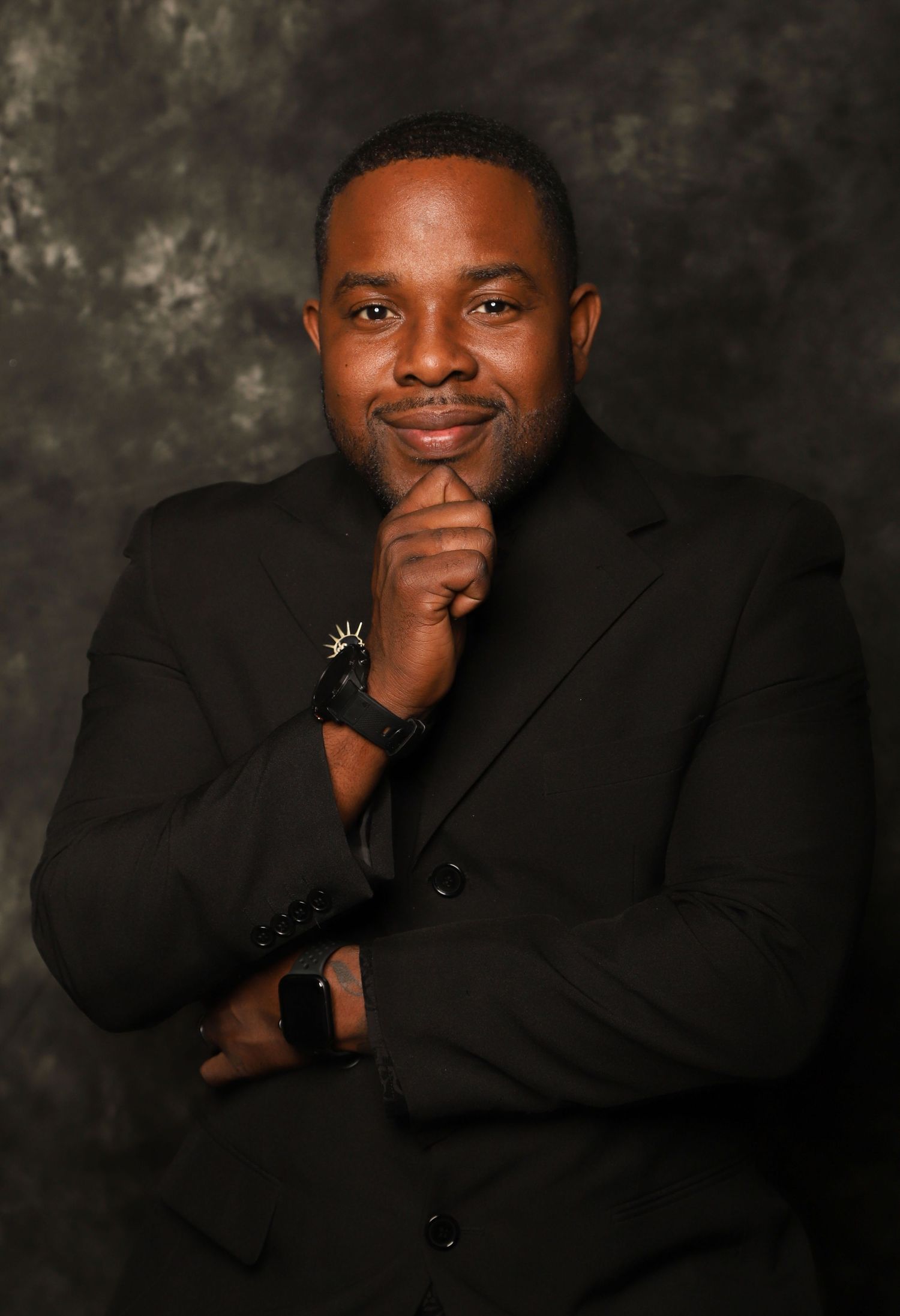 Professional portrait of man in black suit posing confidently with hand on chin in studio portrait.