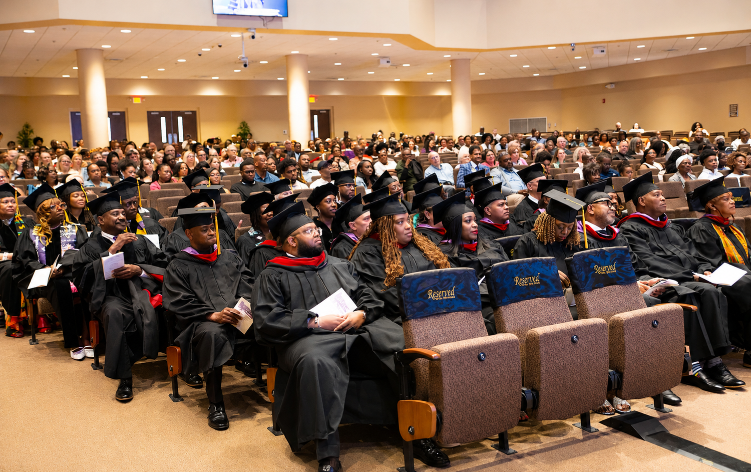 Large graduation ceremony audience seated in academic regalia inside a packed university auditorium.