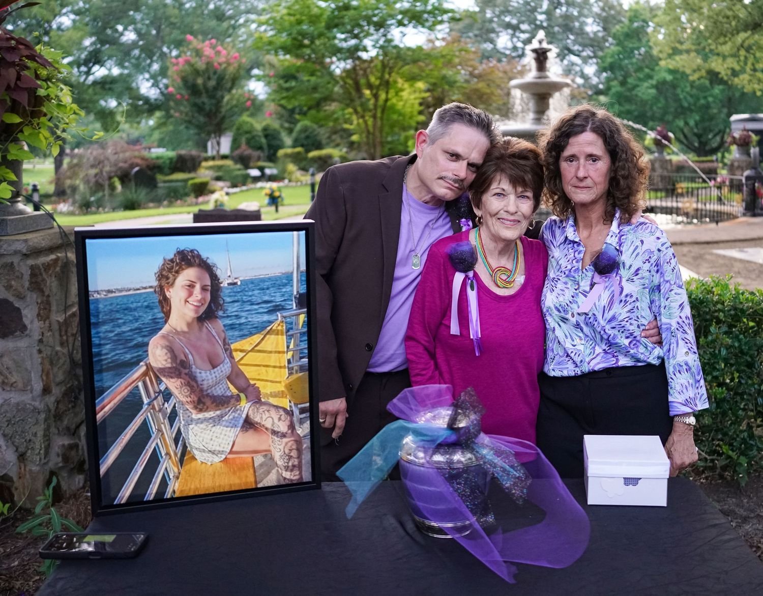 Three people standing together outdoors beside a framed photo and memorial items at a tribute event.