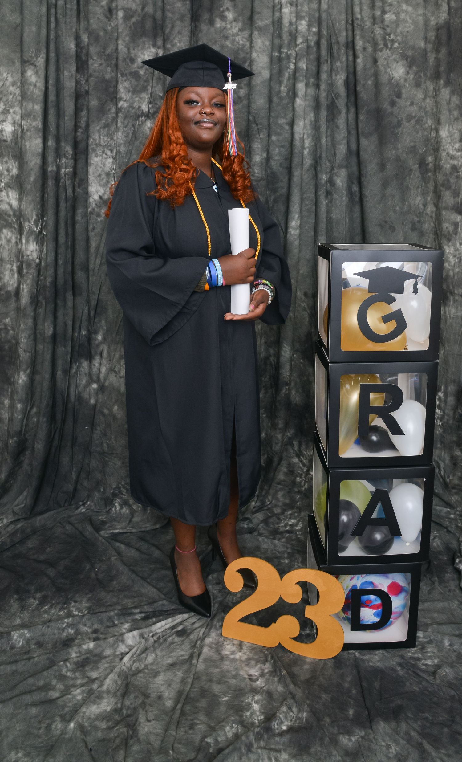 A graduate in black cap and gown poses with GRAD boxes and a 2023 sign.