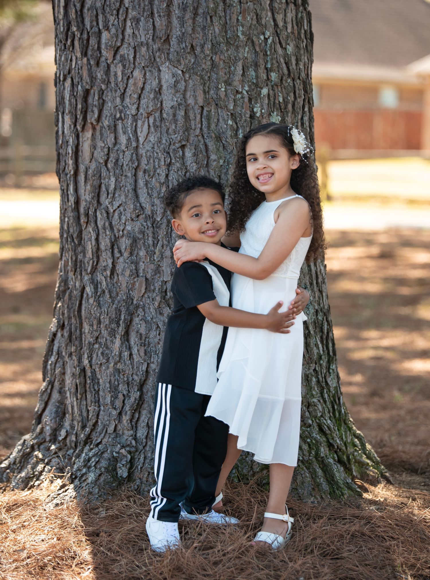 Young boy and girl hugging beside a large tree trunk outdoors.