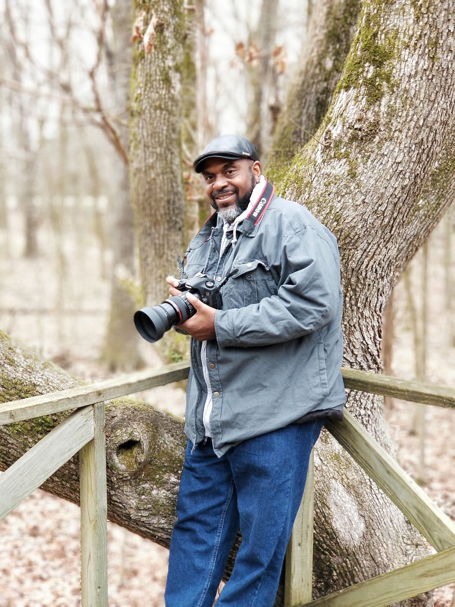 A cheerful photographer poses with his camera beside a wooden fence in a wooded area.
