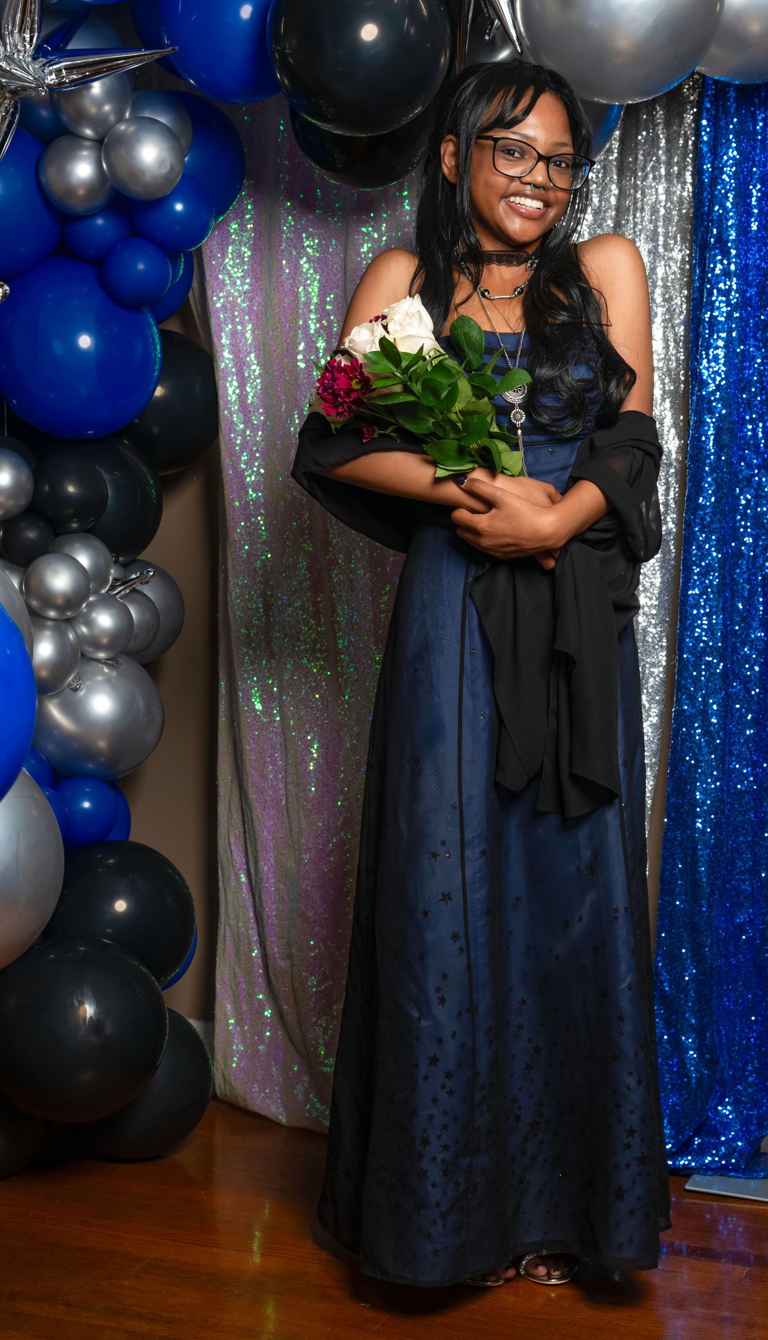 Young woman in navy blue gown holding roses at prom with blue and silver balloon decorations.